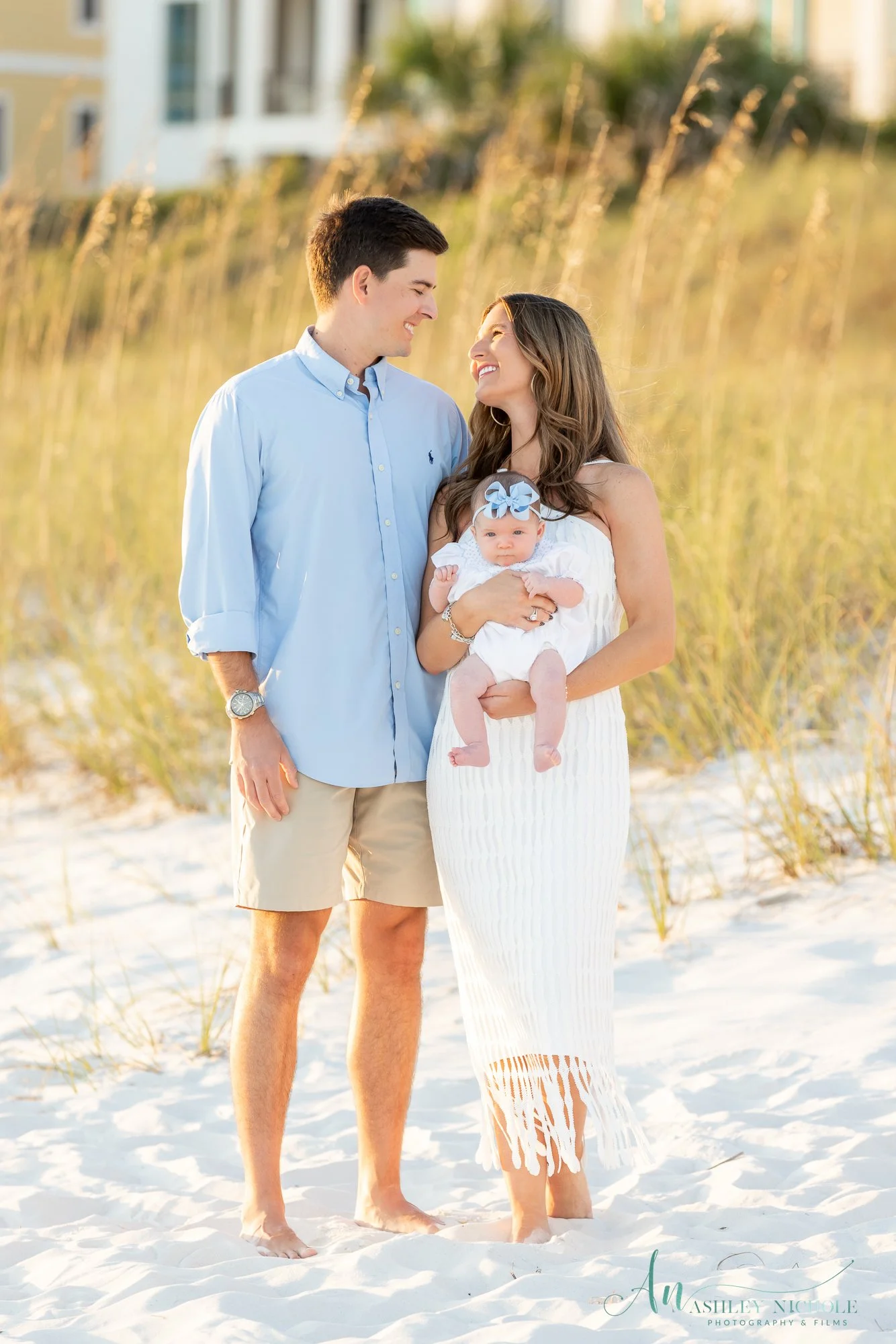 A young family standing on the beach, smiling and looking at each other. The mother holds a baby girl dressed in white with a blue ribbon headband. The father wears a light blue shirt and khaki shorts. The background features tall grass and a buildin