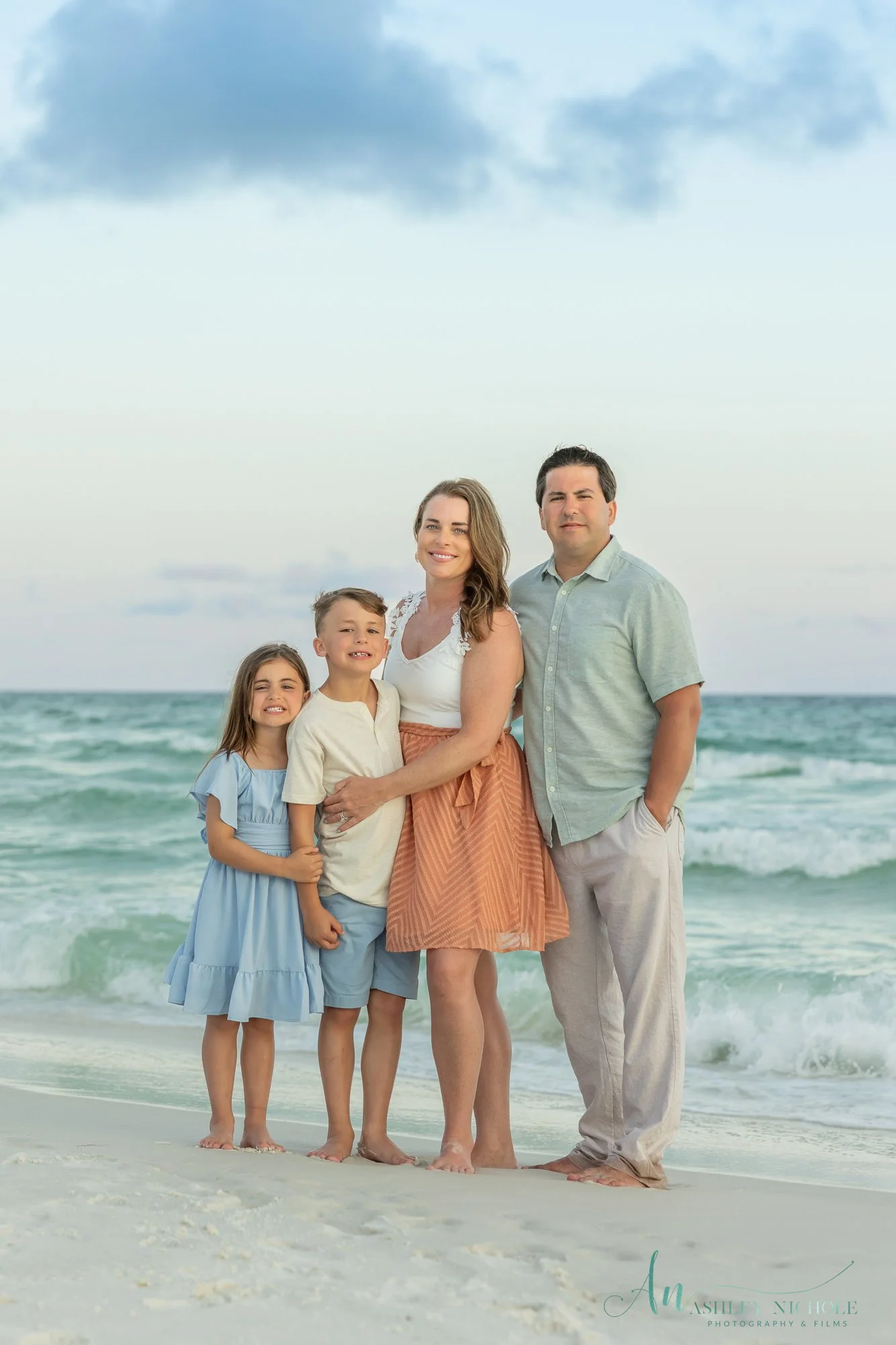 A family of four standing on a sandy beach with the ocean and sky in the background, all smiling and dressed in casual summer clothing.