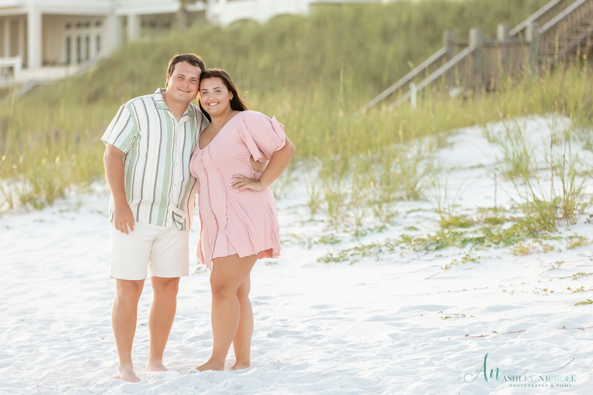 A smiling couple standing on a beach in front of grassy dunes and a house, with the woman wearing a pink dress and the man in a striped shirt and white shorts.