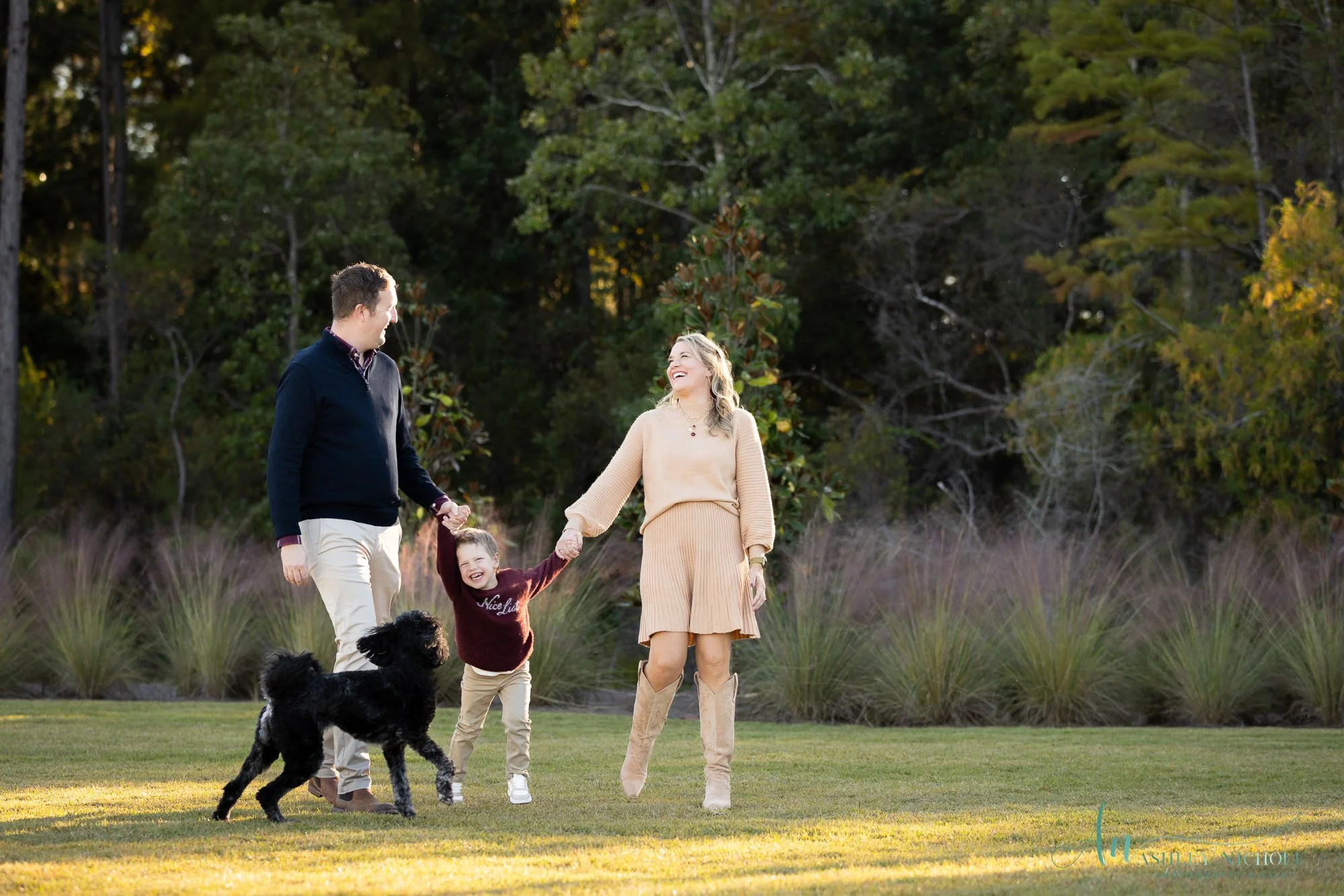 A family of four, including a woman, man, young girl, and dog, enjoying outside on a grassy area with trees in the background. The woman and girl are holding hands, and everyone is smiling.