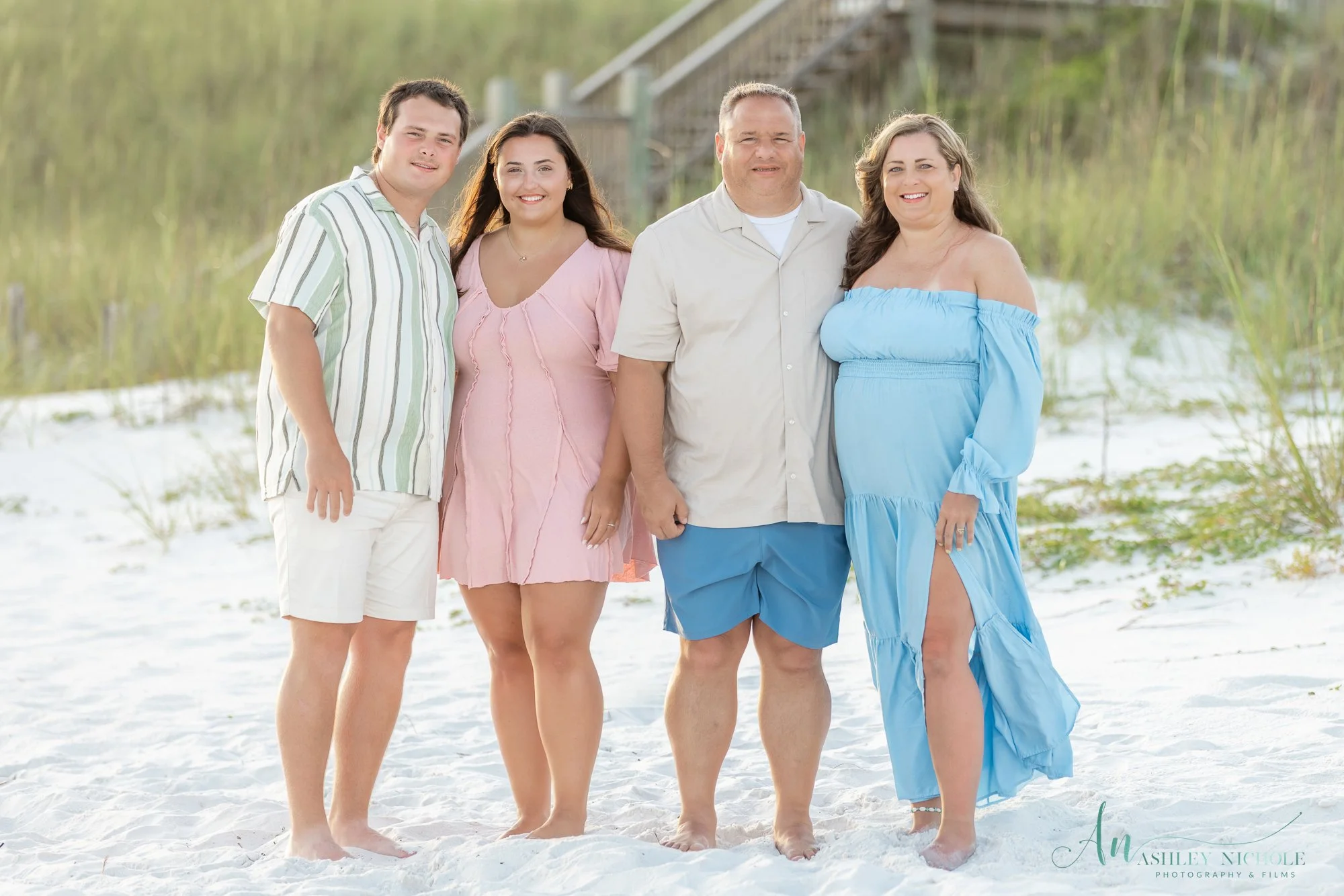 A group of five people standing on a sandy beach with grass and a wooden fence in the background.