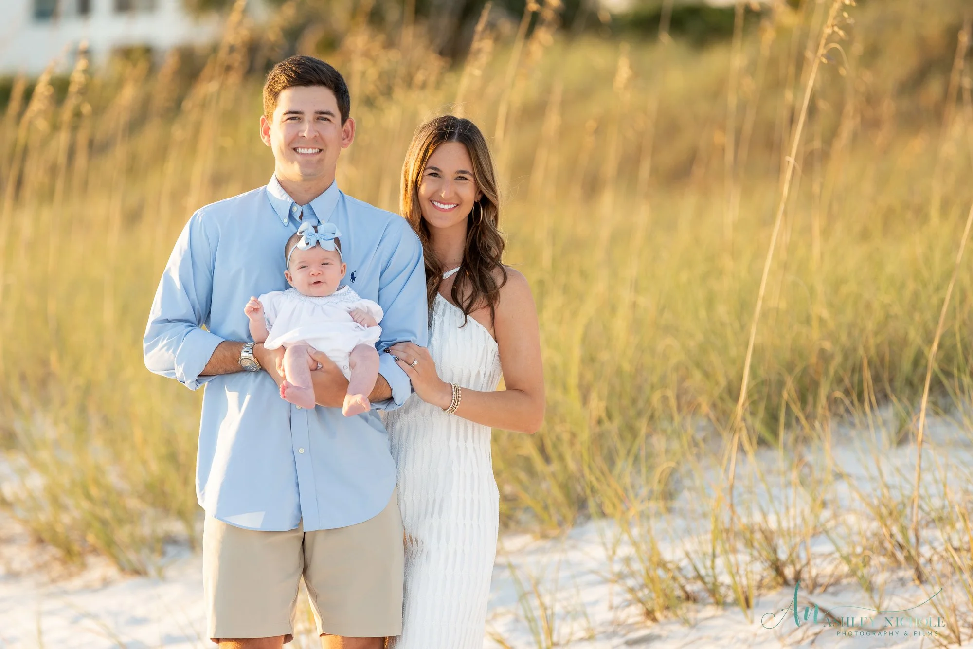 A smiling family of three standing on a sandy beach with tall grass in the background during sunset. The father is holding a baby girl dressed in white with a blue bow. The mother stands beside them, smiling at the camera.