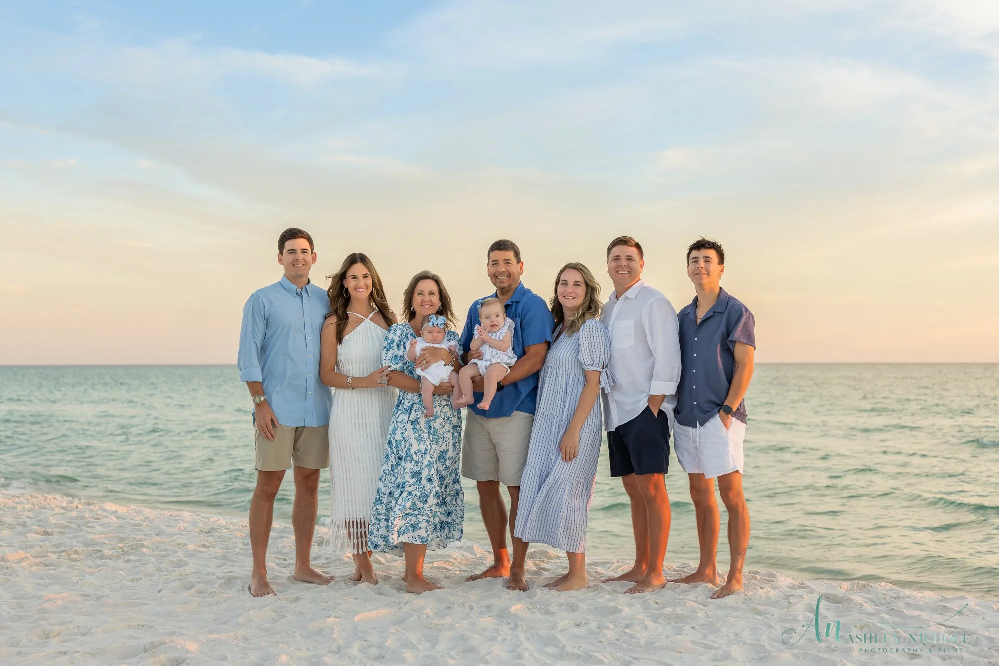 Group of nine people on a beach at sunset, smiling and standing side by side, with the ocean in the background.