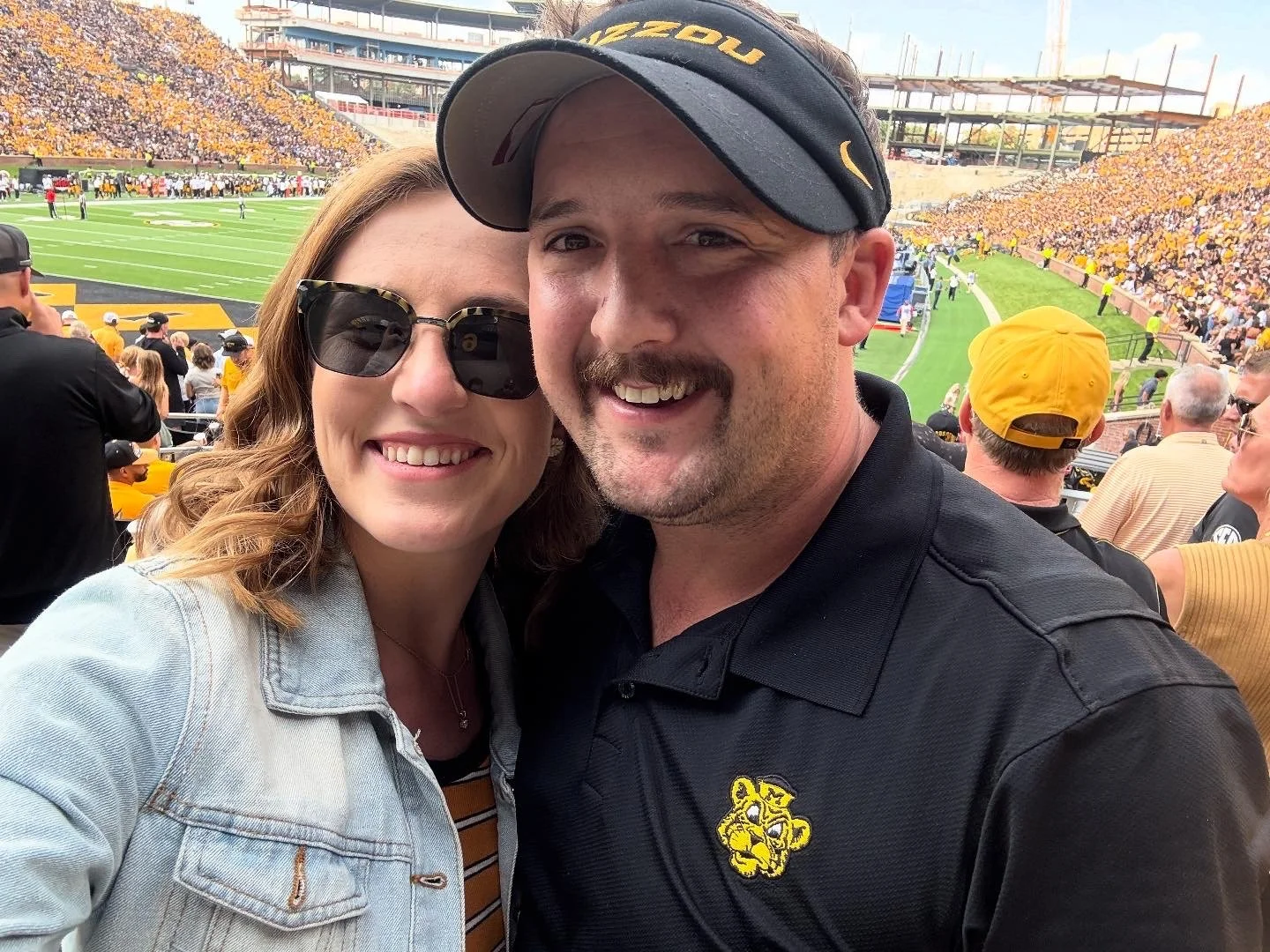 A smiling couple taking a selfie at a football game. The woman is wearing sunglasses and a denim jacket, the man is wearing a black shirt with a sports logo and a cap. The stadium is filled with fans, and the football field is visible in the background.