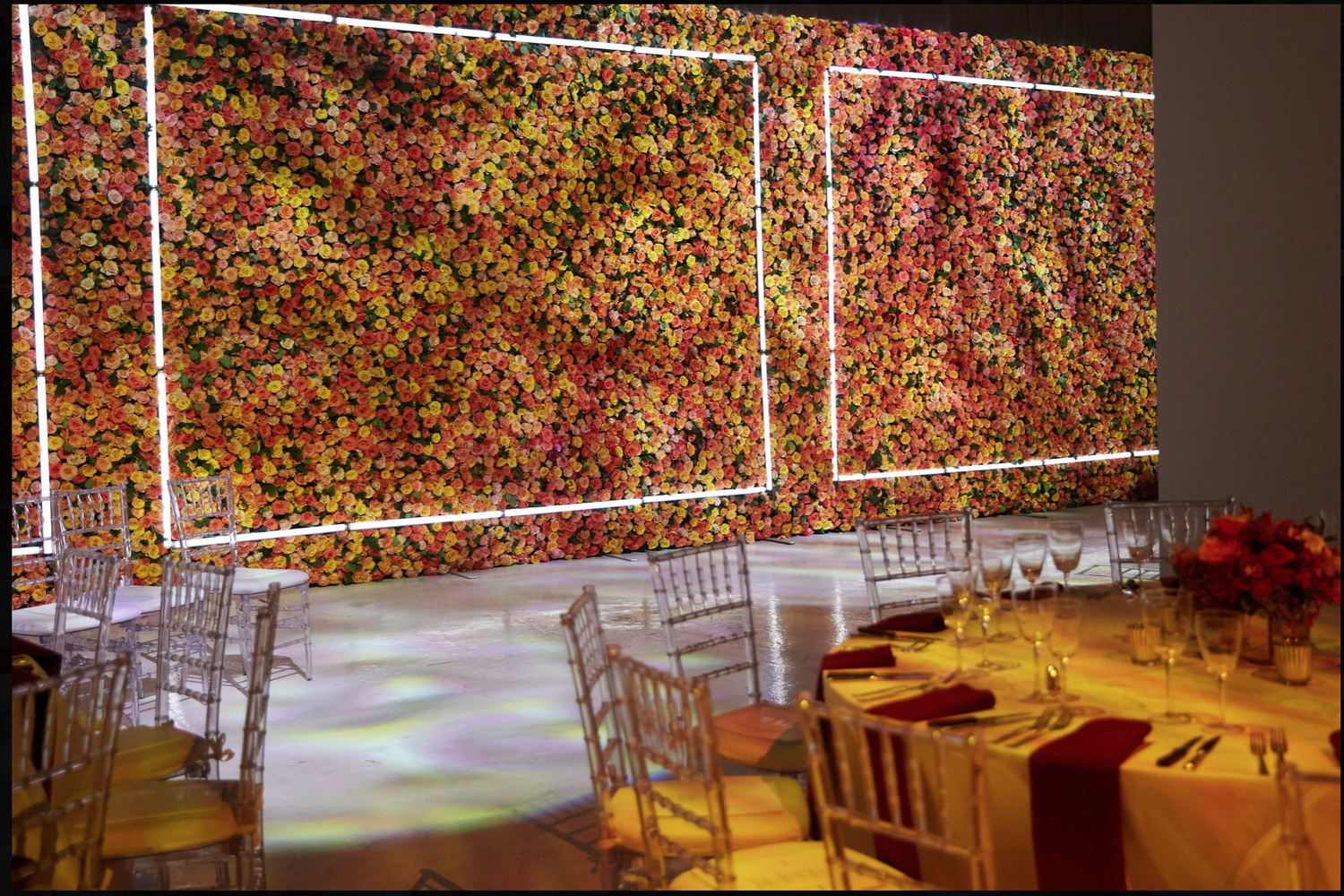 A wall of red, orange and yellow flowers framed by glowing light panels at an evening event