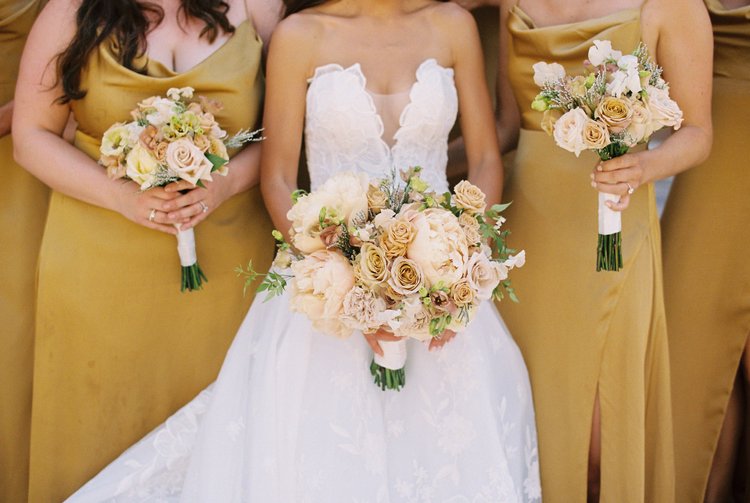 A bride holding a bouquet of blush and cream roses beside bridesmaids in gold