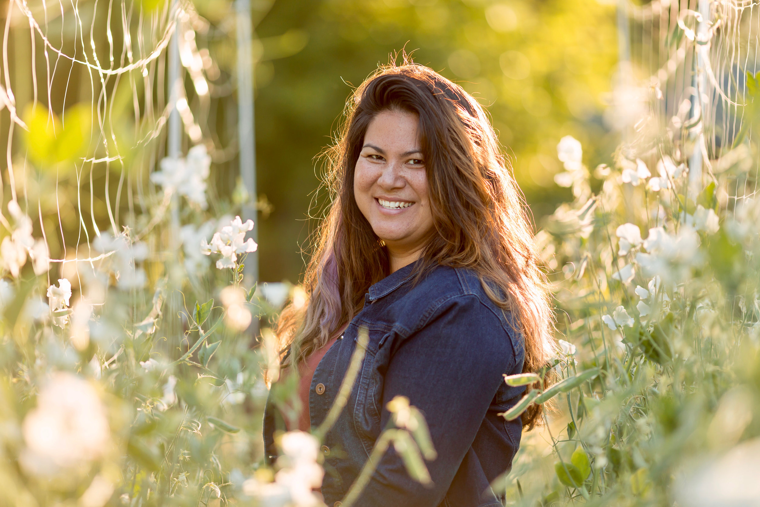 ashley at golden hour in a row of sweet pea flowers, female with long brown hair and light purple at the ends in the center of the picture wearing a dark blue jean jacket and burgundy red top.