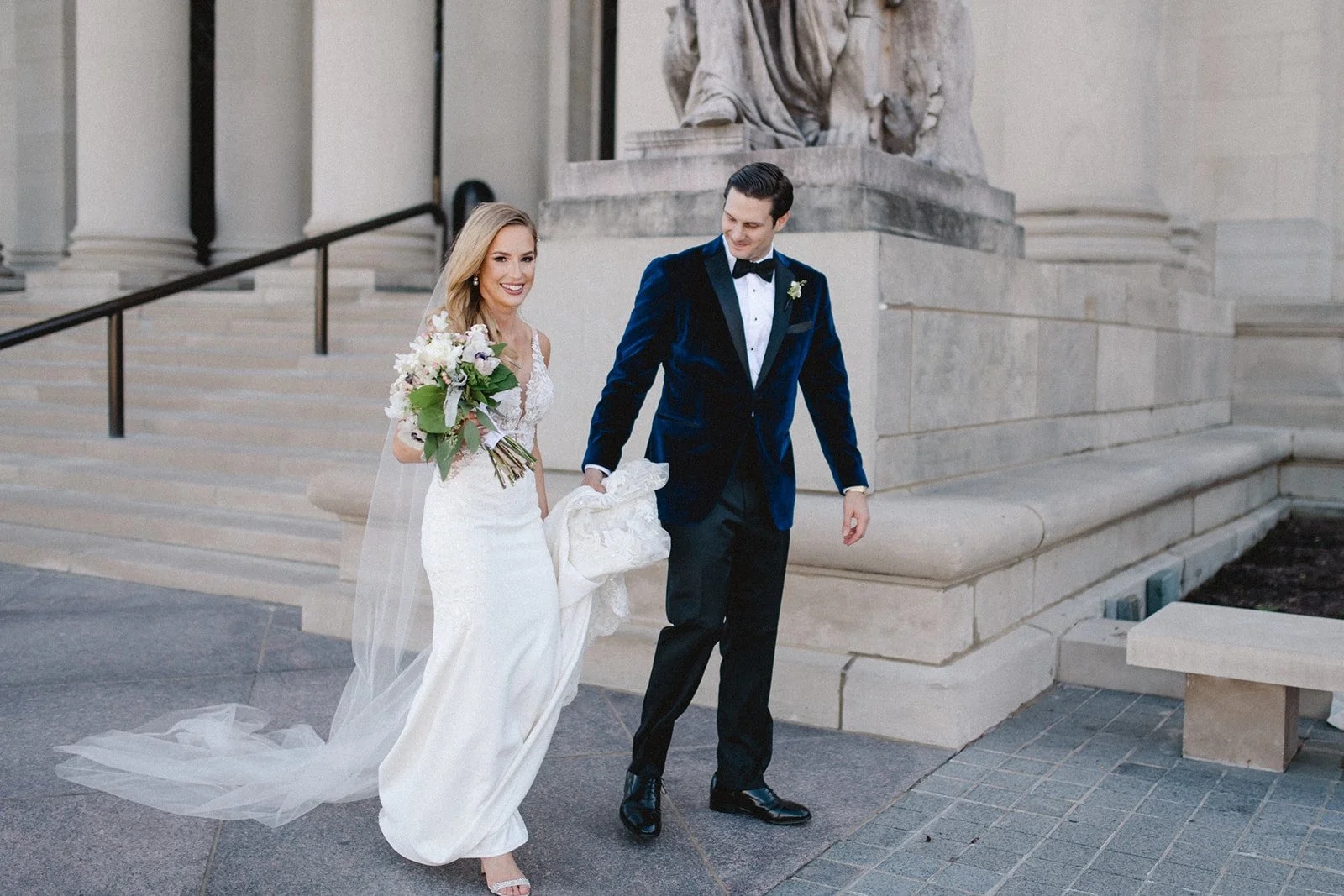 Bride and groom walking together outside of museum, groom holding brides train in his blue suede jacket.