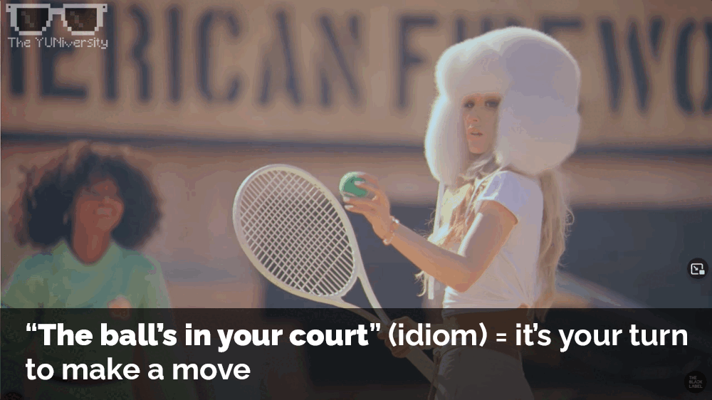 A woman with large white hair styled in a vintage 1960s fashion holding a tennis ball and racquet on a tennis court with a woman smiling in the background. A sign with partially visible text reads 'AMERICAN FU...'.