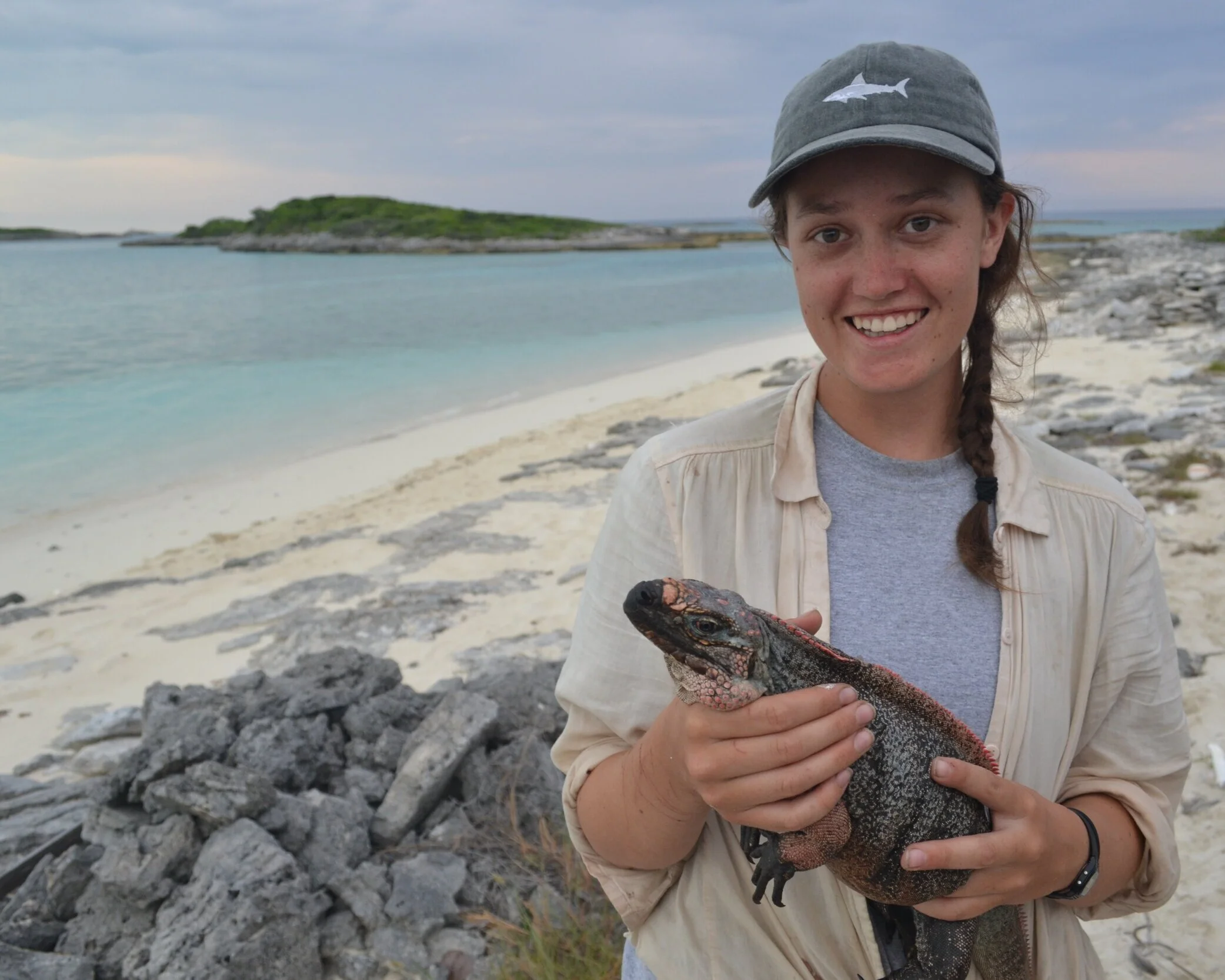 Erin is holding a marine iguana. She’s wearing a baseball hat and is standing on a tropical beach