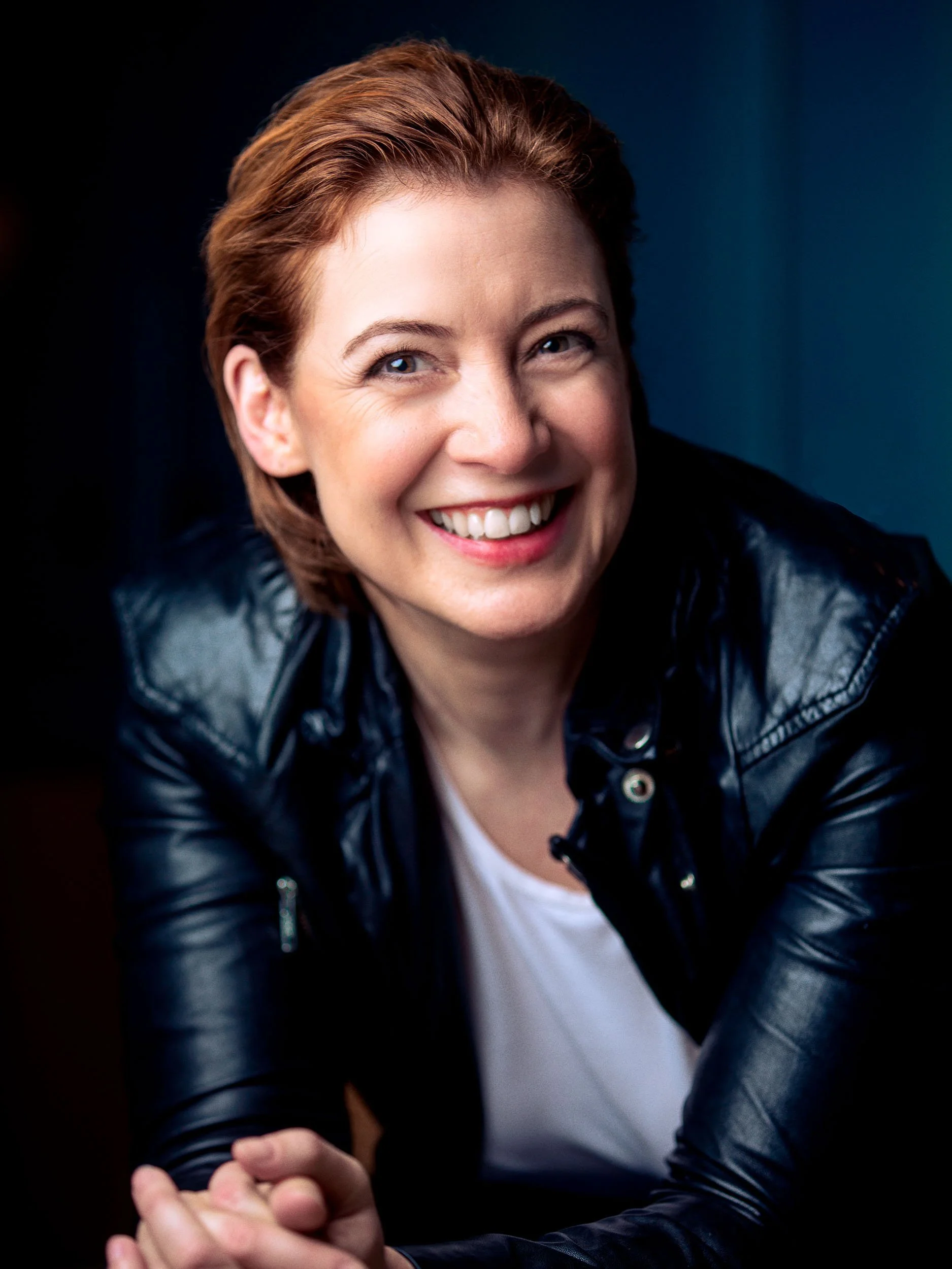 Lachende Frau in schwarzer Lederjacke vor dunklem Hintergrund.