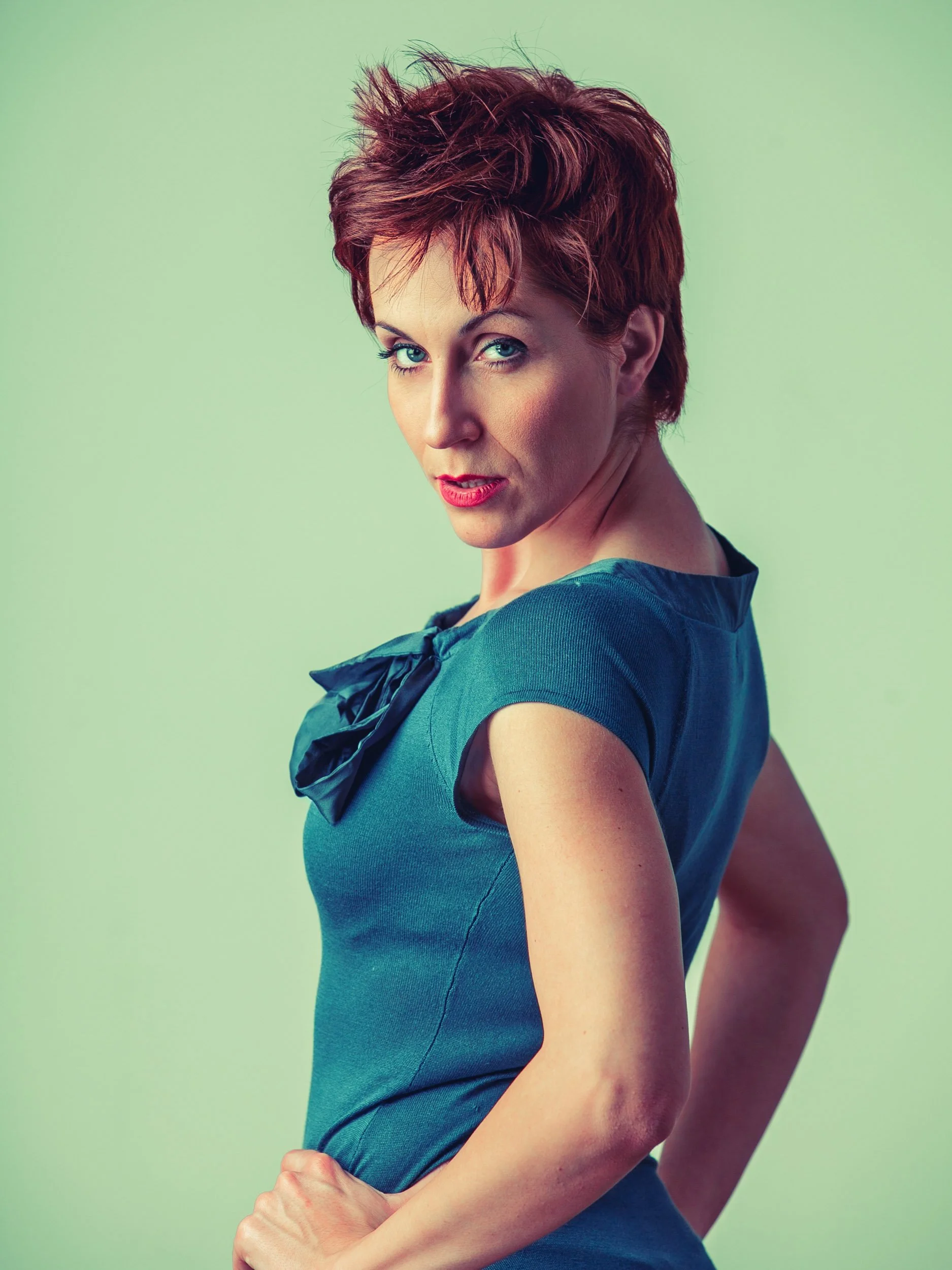 Eine Frau mit kurzen, rötlichen Haaren trägt ein blaues Kleid und schaut über die Schulter zur Kamera. Hintergrund ist einfarbig grünlich.