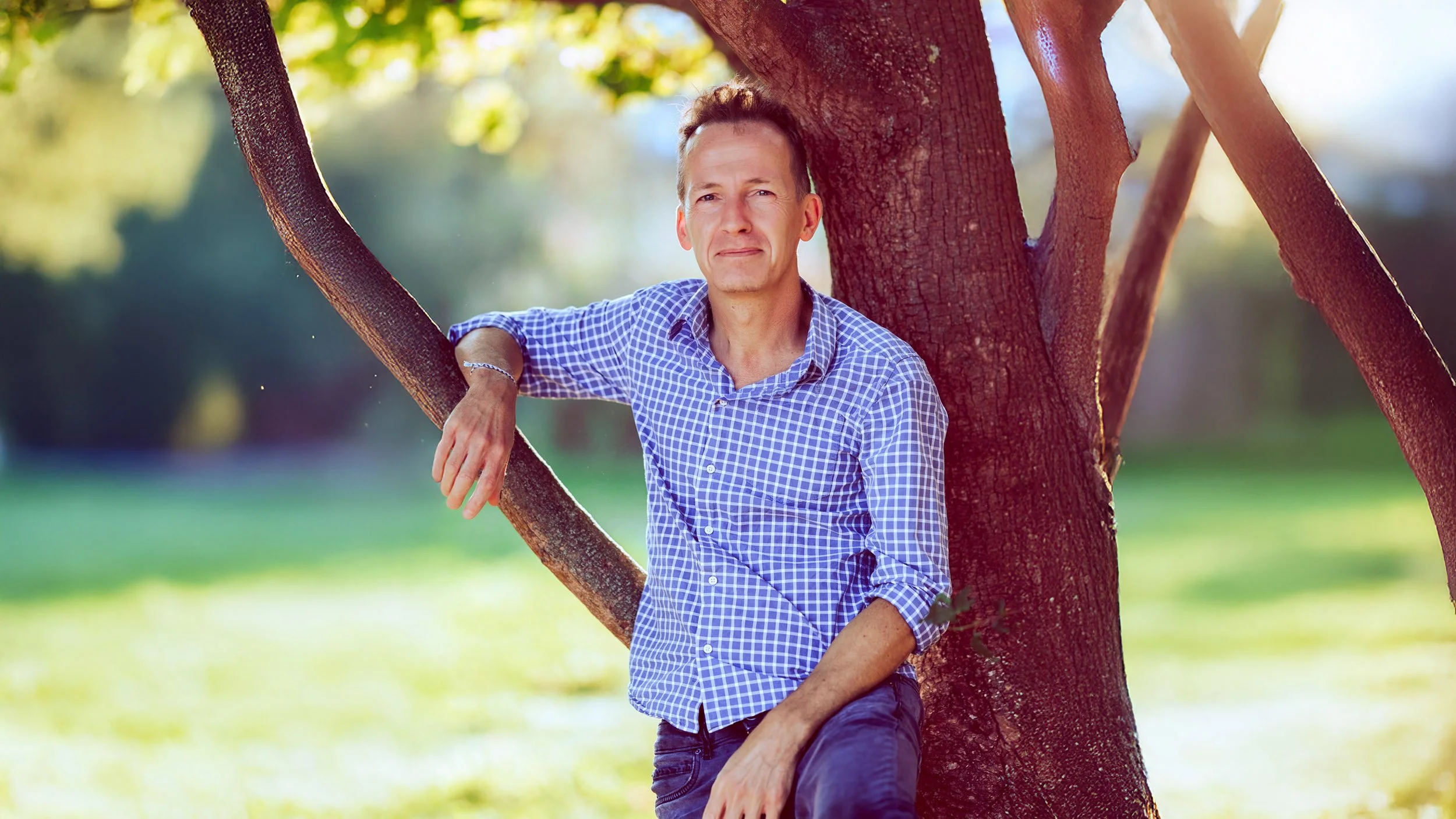 Ein Mann in einem karierten Hemd lehnt an einem Baum in einem sonnigen Park.