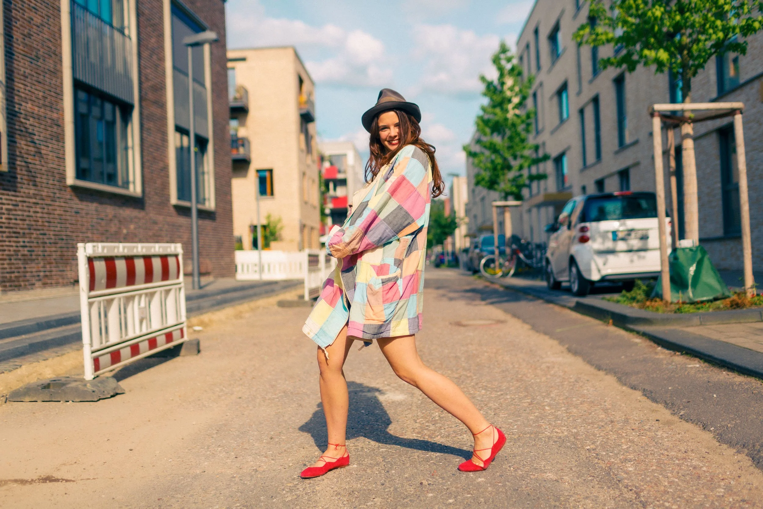 Frau in bunter Kleidung und rotem Schuhen geht auf einer städtischen Straße, umgeben von modernen Gebäuden und parkenden Autos.