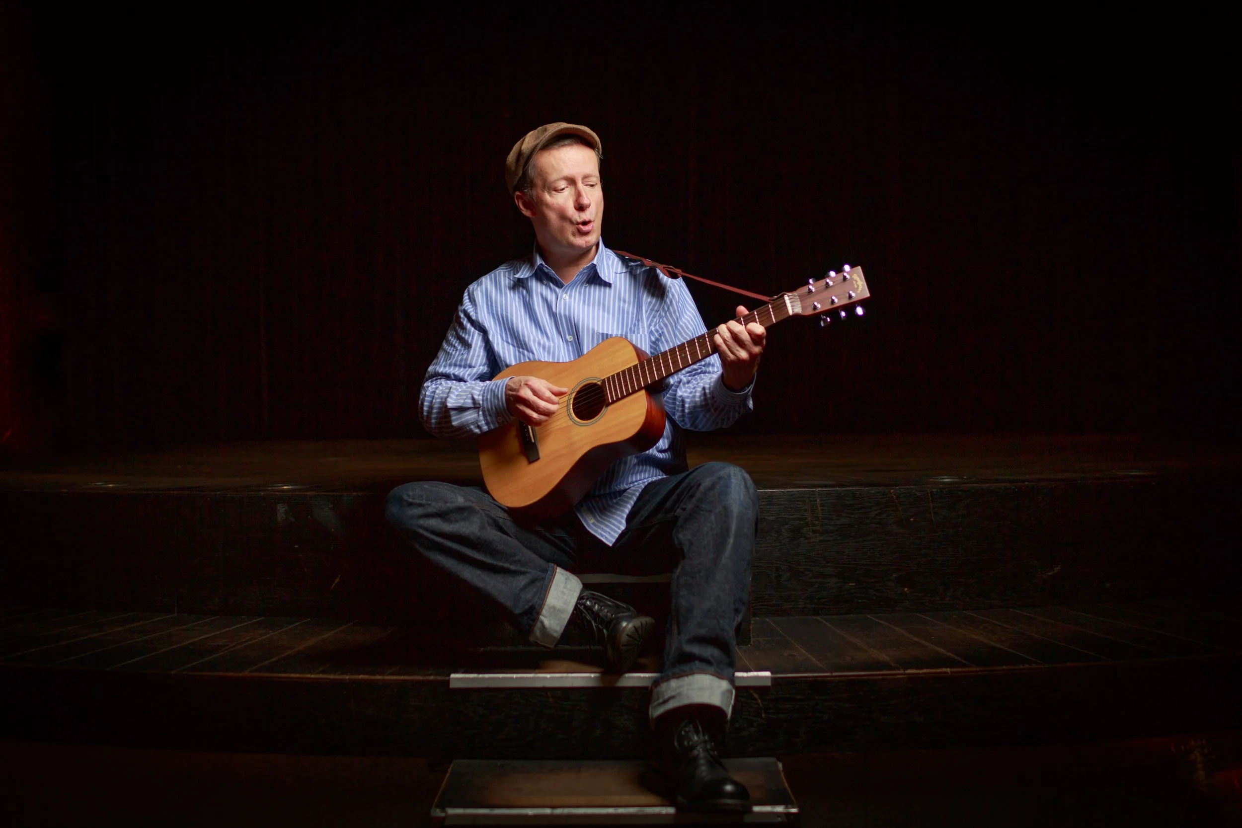 Ein Mann sitzt auf einer dunklen Bühne, spielt Gitarre und trägt eine blaue gestreifte Hemd und eine Mütze.