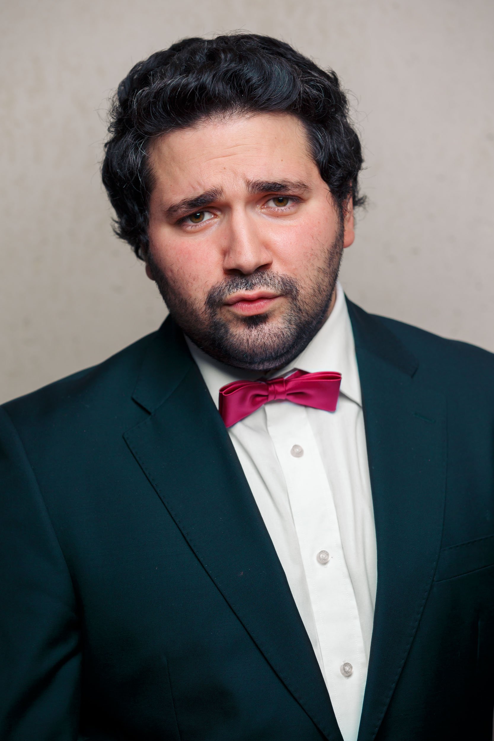 Ein Mann in einem schwarzen Anzug mit einem weißen Hemd und einer roten Fliege, neutraler Gesichtsausdruck, vor einem beigen Hintergrund.