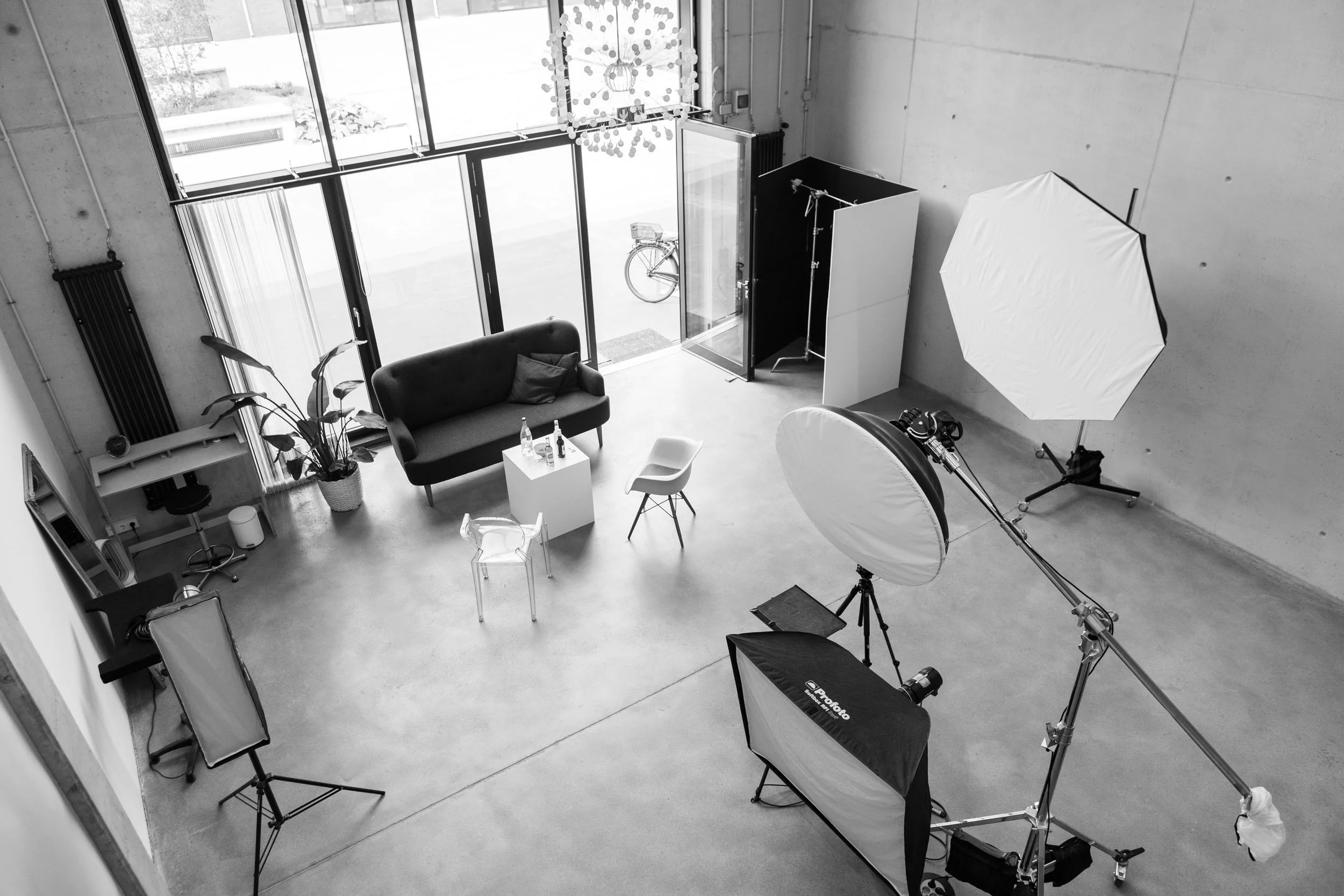 Fotostudio mit professionaler Beleuchtung, Sofa, Tisch mit Getränken, Fahrrad vor Fenster, Pflanzen, und Fotografieausrüstung.