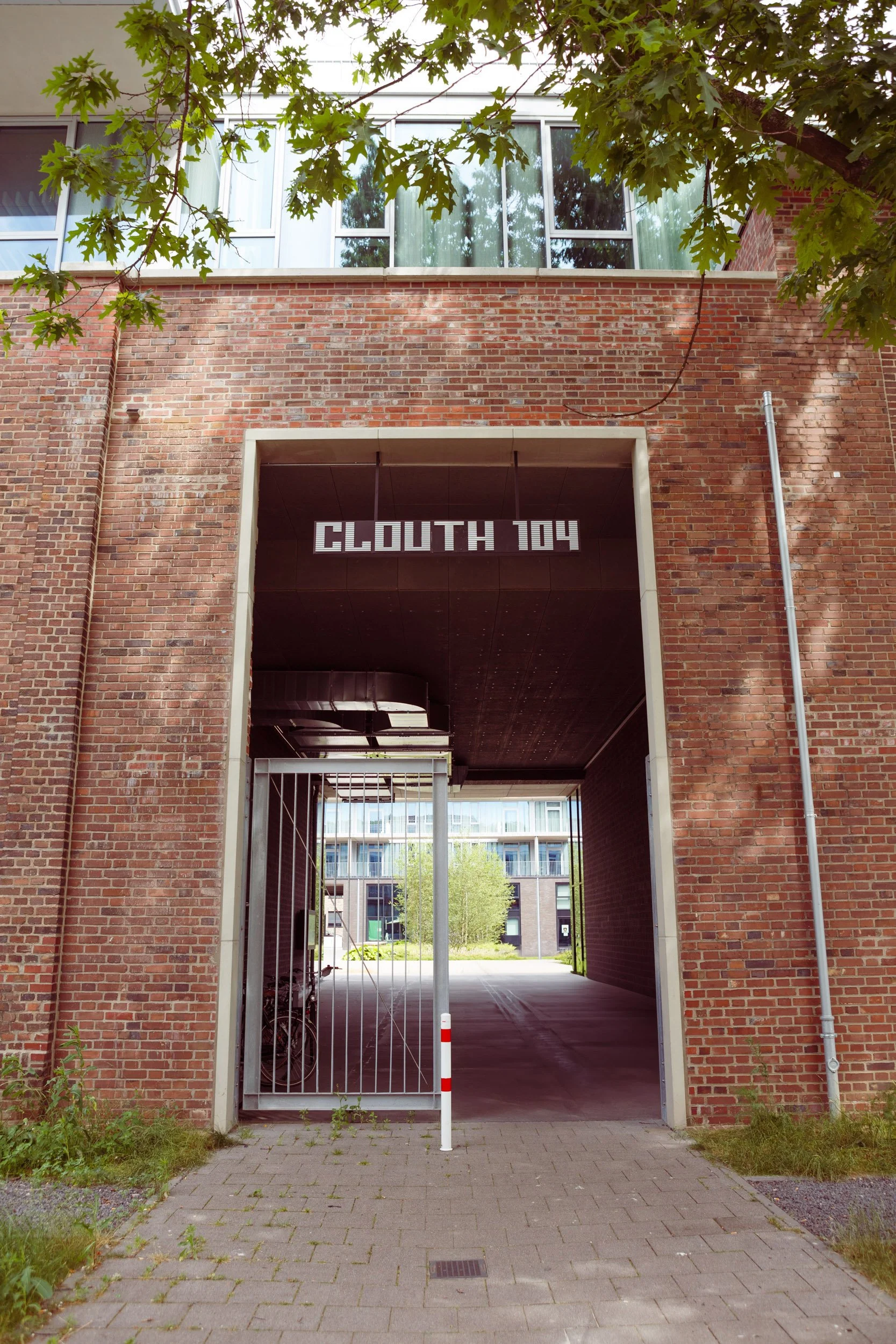 Der Eingang eines modernen Gebäudes mit einem roten Ziegelstein-Facade und einem metallischen Gittertor. Über dem Tor ist ein digitales Schild mit der Aufschrift "Clouth 104". Im Hintergrund sind Wohnungen und Bäume sichtbar.