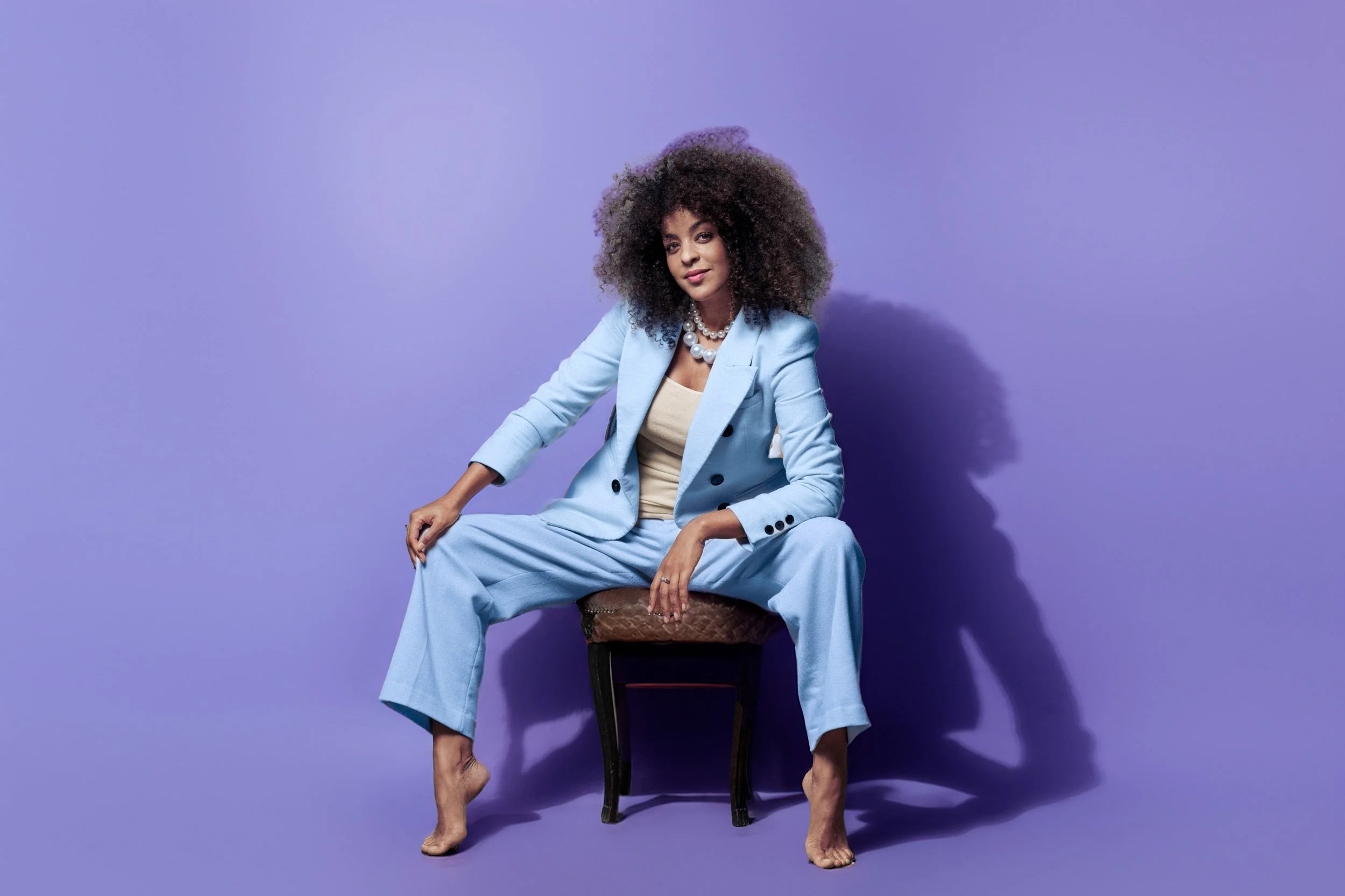 Eine Person in einem hellblauen Anzug sitzt barfuß auf einem Stuhl vor einem lila Hintergrund.