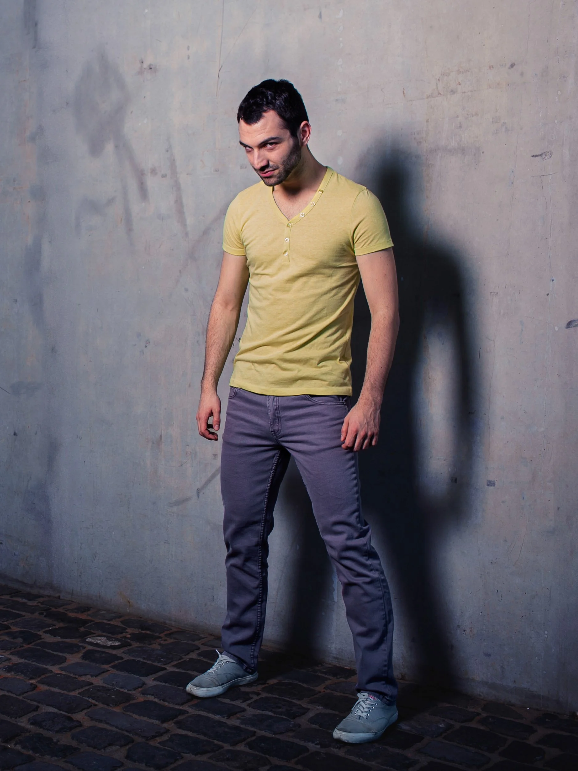 Mann in gelbem T-Shirt und grauer Hose steht vor einer Betonwand.