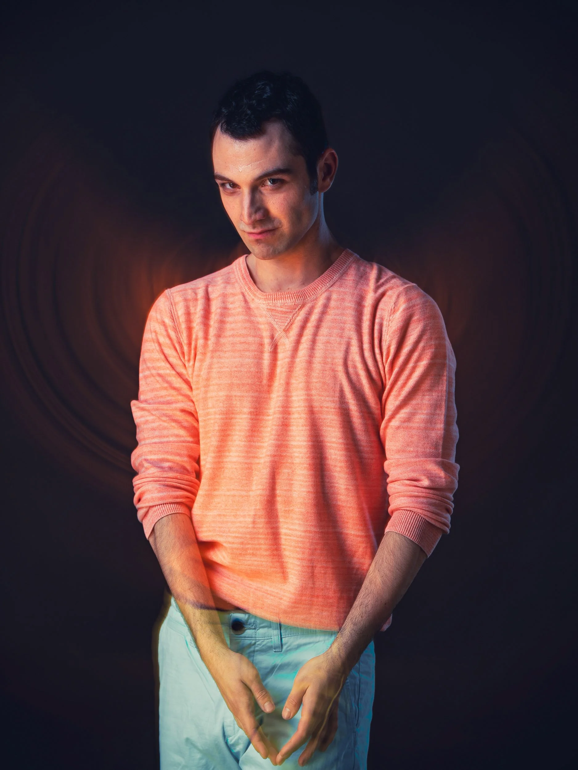 Ein Mann in einem orangefarbenen Pullover und hellblauer Hose posiert vor einem dunklen Hintergrund.