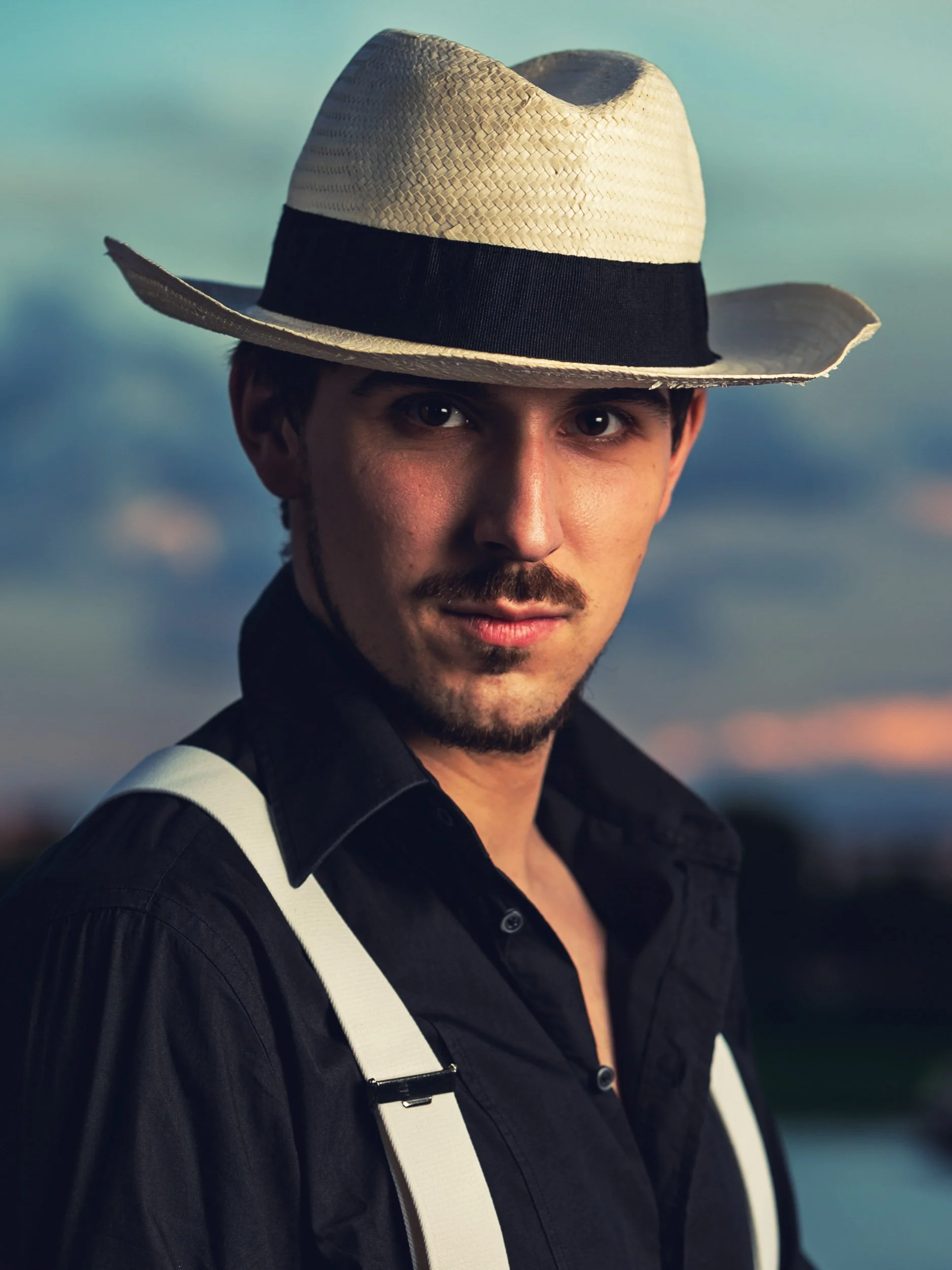 Mann mit Strohhut und Hosenträgern, trägt ein schwarzes Hemd. Hintergrund leicht unscharf in Dämmerungsfarben.