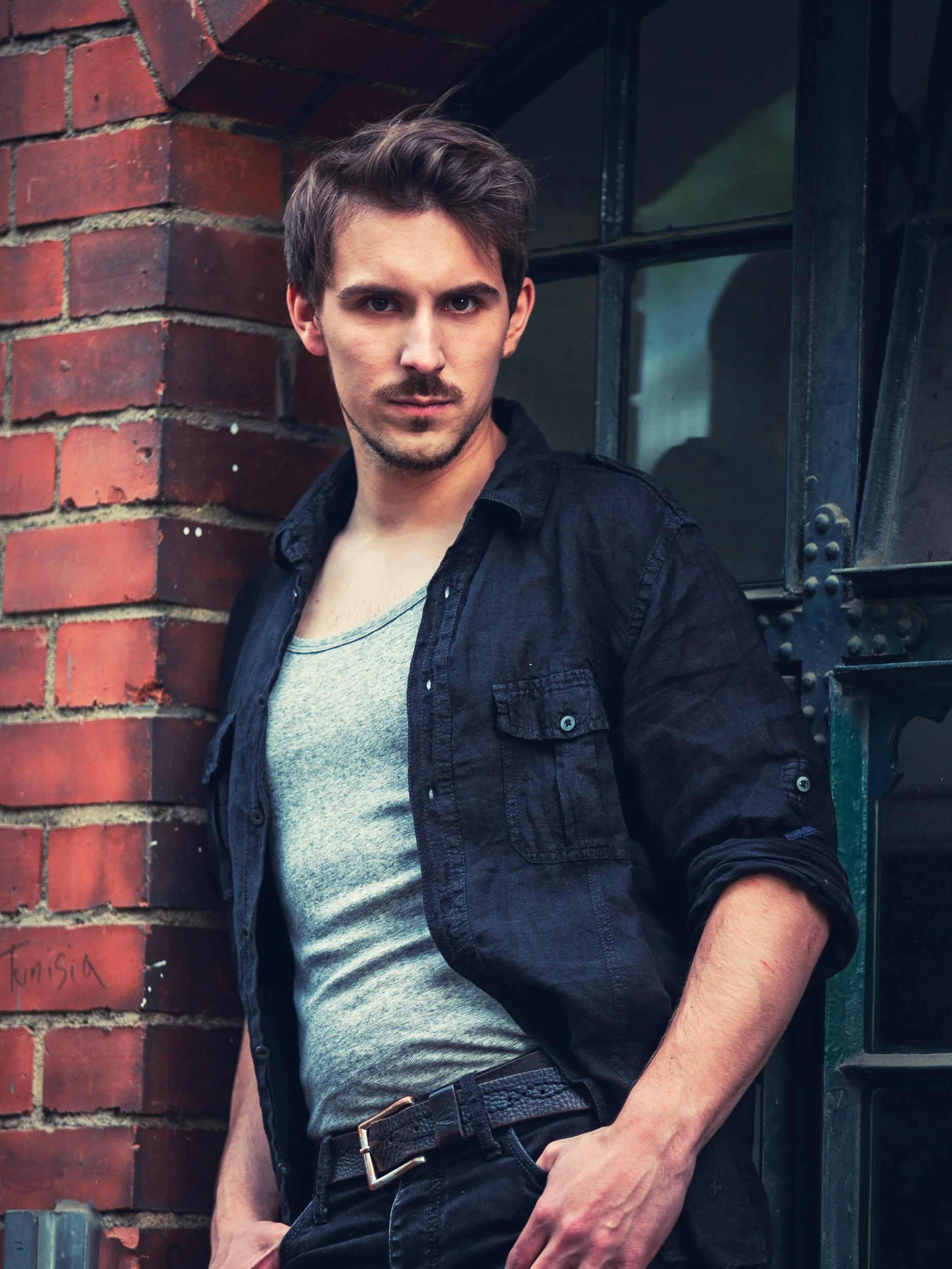 Mann in grauem Tanktop und schwarzem Hemd lehnt an einer Ziegelwand, neben einem Fenster, schaut direkt in die Kamera.