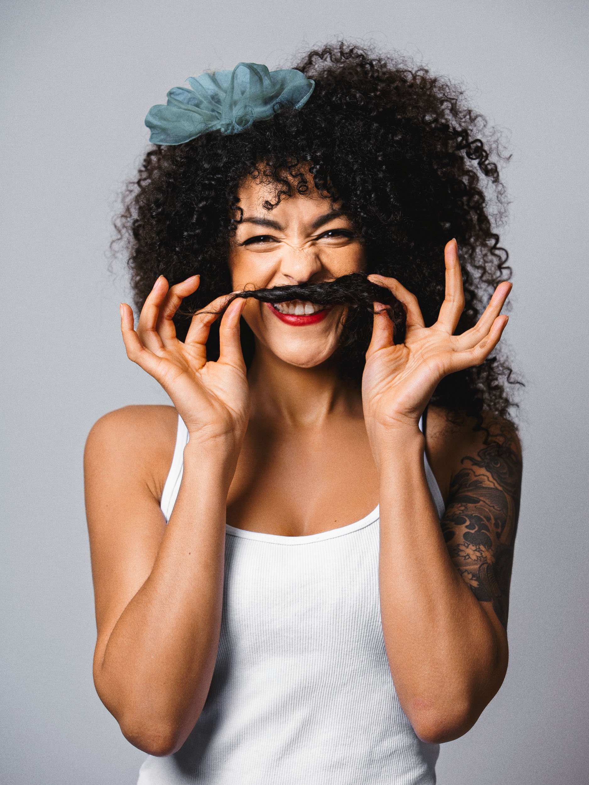 Frau mit lockigem Haar und blauer Schleife formt mit ihrem Haar einen Schnurrbart, lächelt, trägt ein weißes Oberteil.