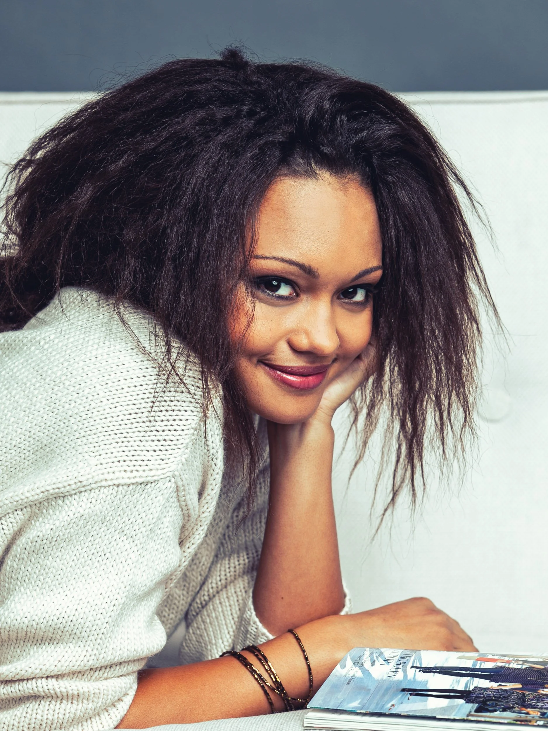 Frau in weißem Pullover, stützt Kopf auf Hand und lächelt, mit Zeitschrift vor ihr liegend.