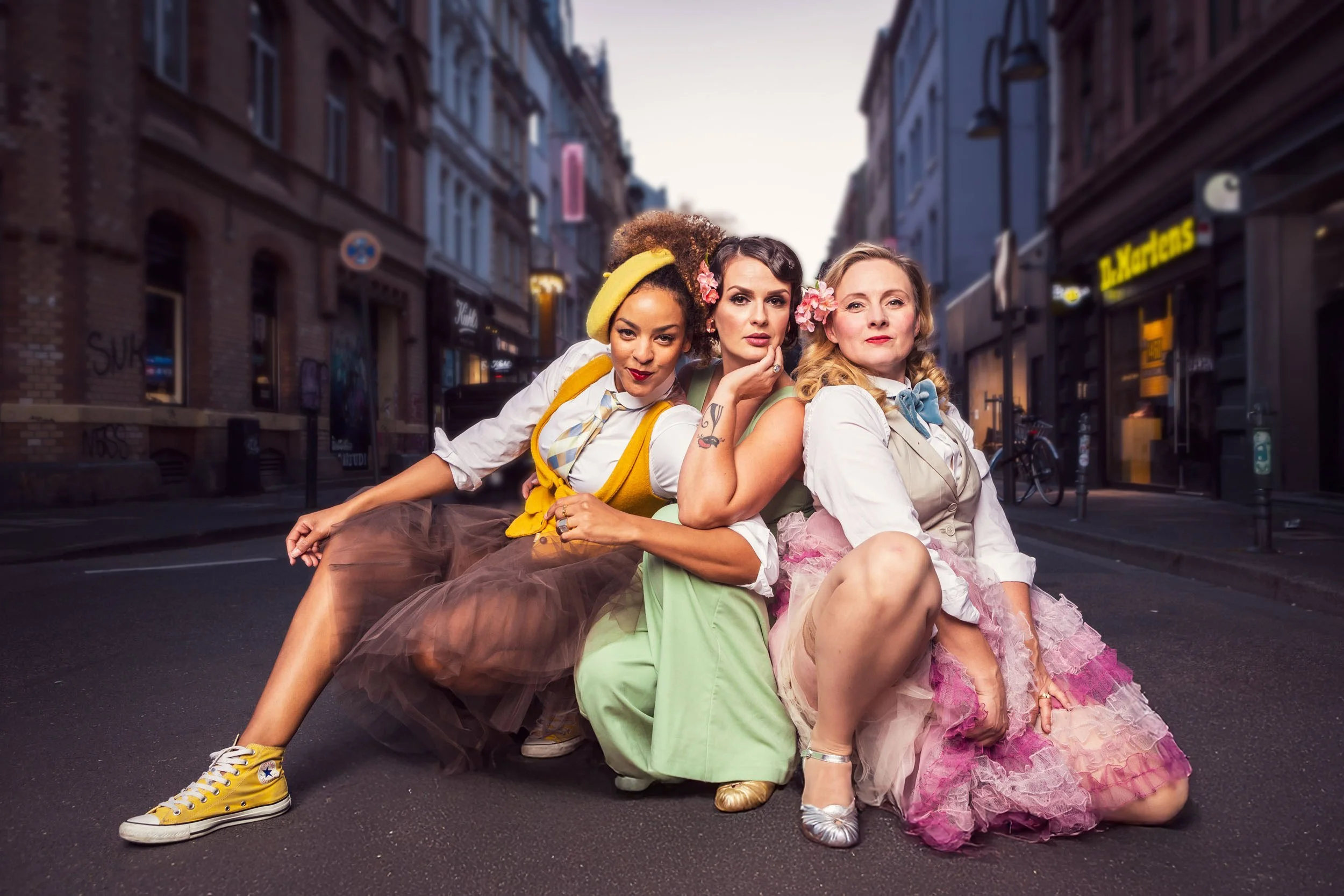 Drei Frauen, stilvoll gekleidet, posieren auf einer städtischen Straße. Die Straße ist von Altbauten gesäumt. Eine Frau trägt gelbe Sneakers, eine andere einen tannengrünen Jumpsuit und die dritte einen pinken Rock. Die Atmosphäre ist verspielt und modisch.