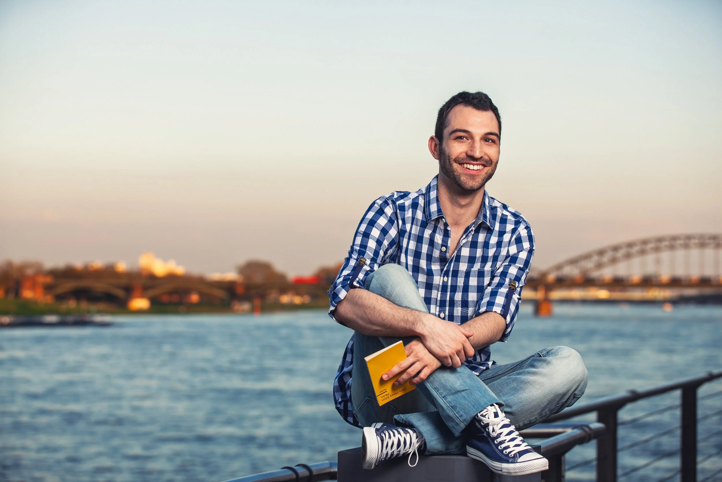 Mann in kariertem Hemd sitzt lachend am Flussufer mit Buch in der Hand, Brücke im Hintergrund.