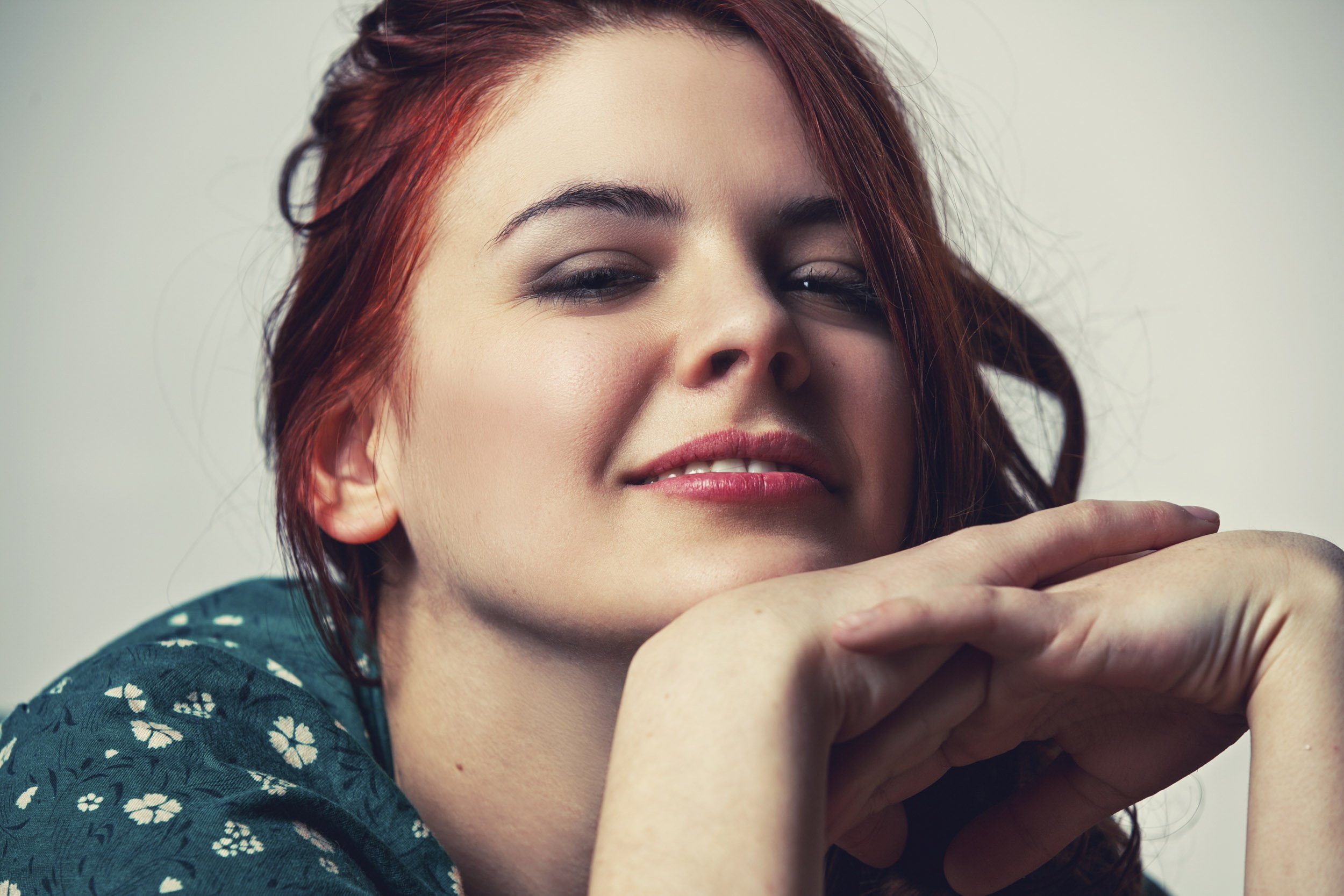 Nahaufnahme einer lächelnden Frau mit roten Haaren und einem grünen Oberteil. Ihre Hände sind unter ihrem Kinn gefaltet.