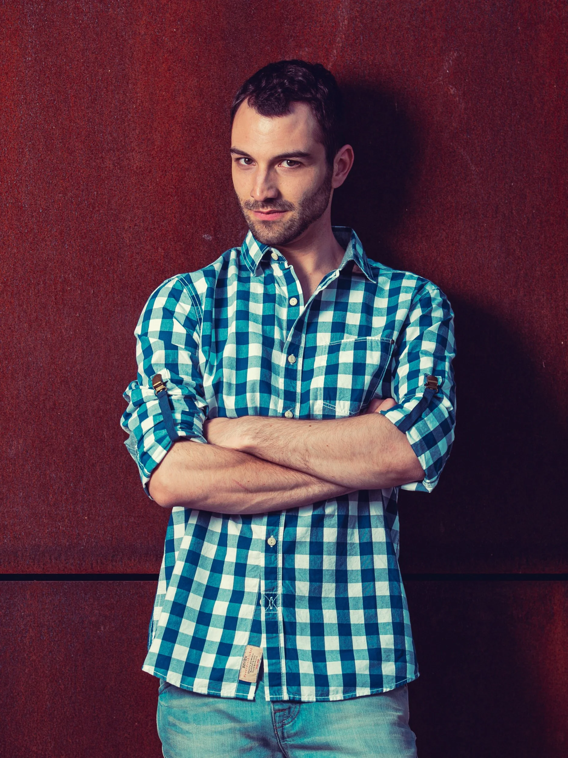 Mann in kariertem Hemd mit verschränkten Armen vor rostrotem Hintergrund.
