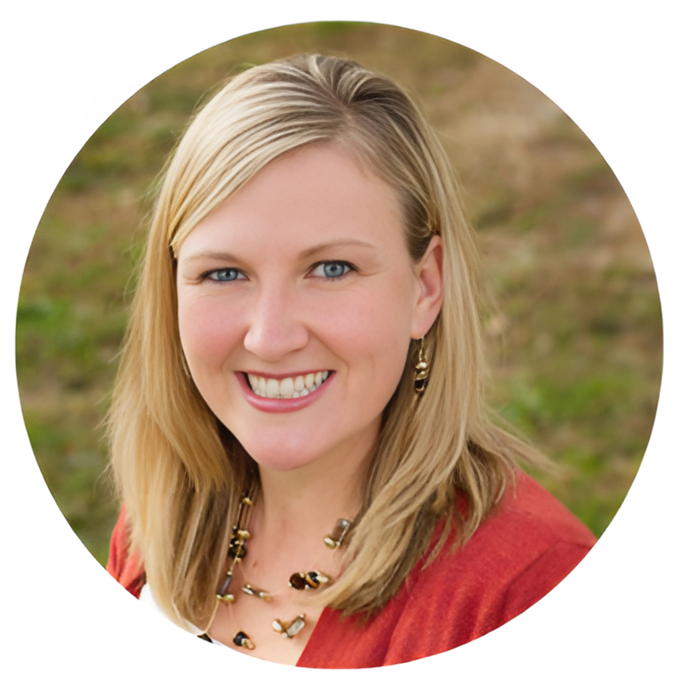 A woman with blonde hair smiling outdoors, wearing a red top and jewelry, with a blurred natural background.