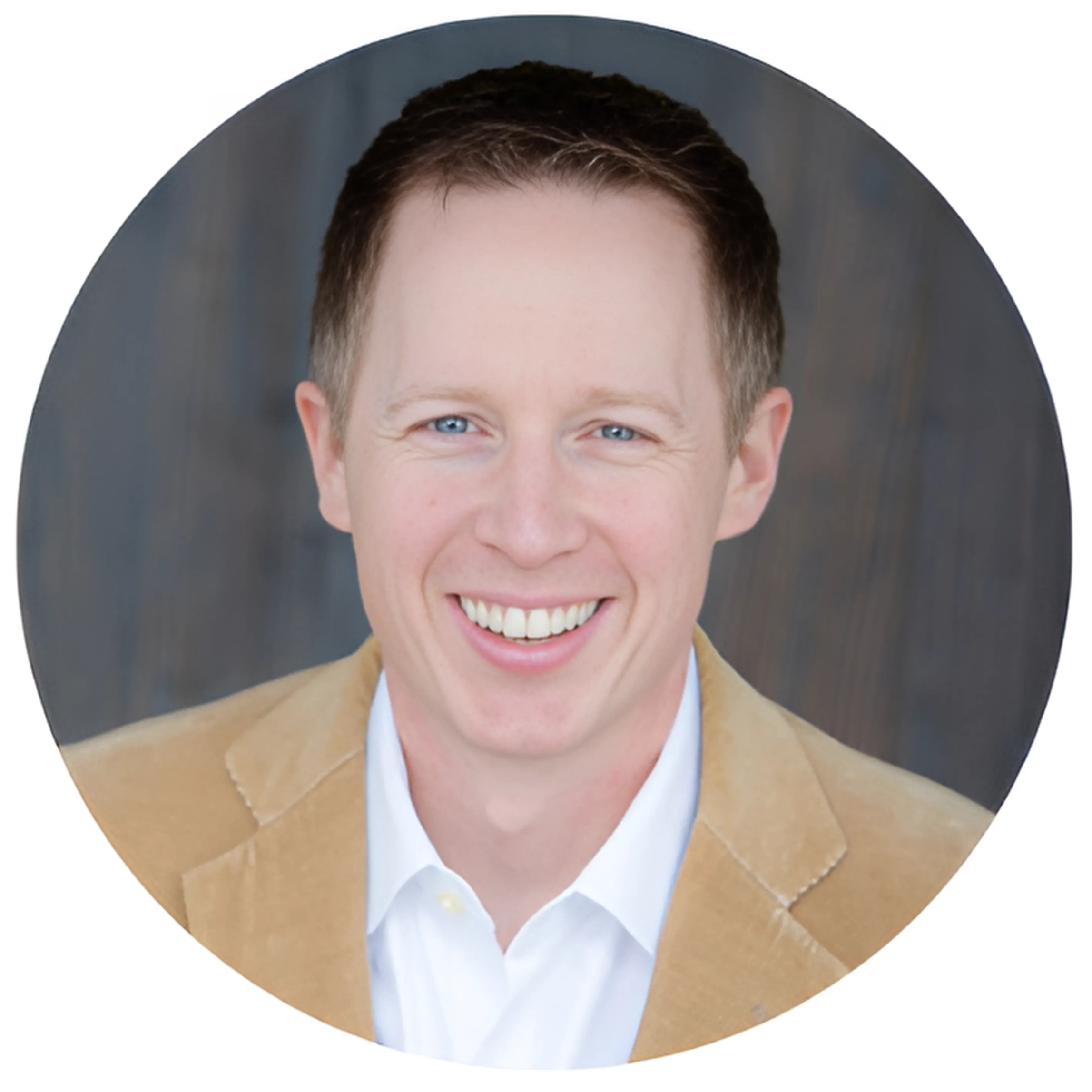 Headshot of a smiling man with short brown hair, blue eyes, wearing a tan suit jacket and white shirt, against a blurred dark background.