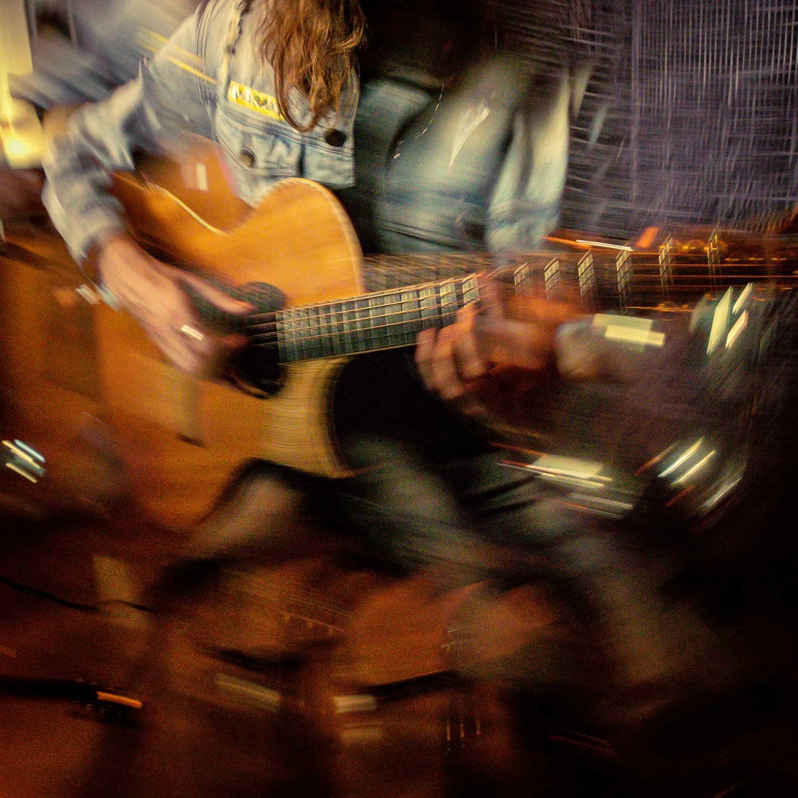 Blurred image of a person playing an acoustic guitar, wearing a striped denim jacket, with a dark background featuring a building or wall with a pattern of lines.