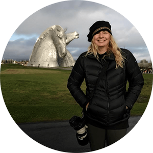 A woman smiling outdoors in front of the Kelpies, large horse head sculptures, in Scotland. She is wearing a black beanie and jacket, holding a camera.