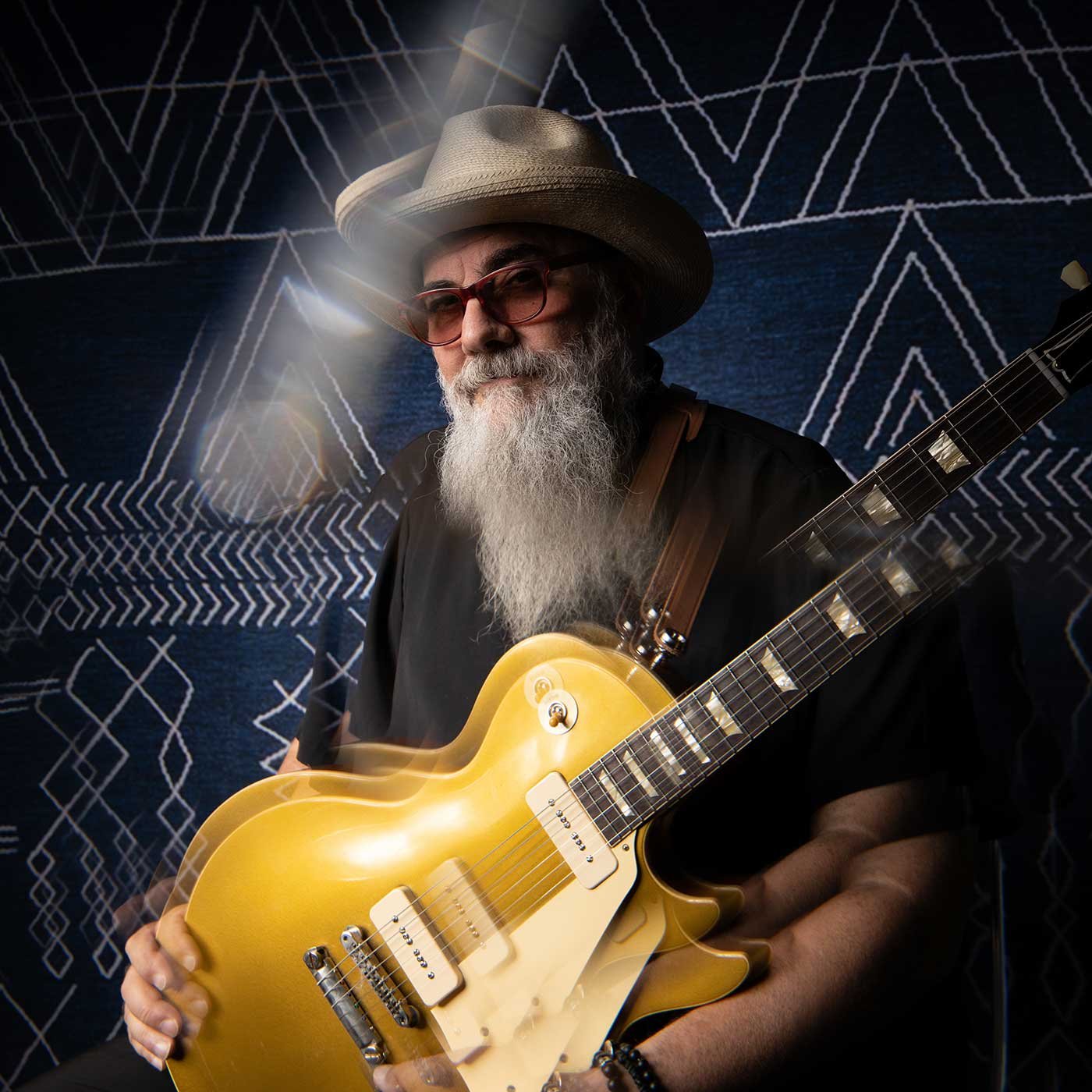 An elderly man with a long white beard and mustache, wearing glasses, a hat, and a black shirt, playing a yellow electric guitar. The background features dark blue fabric with white geometric patterns.