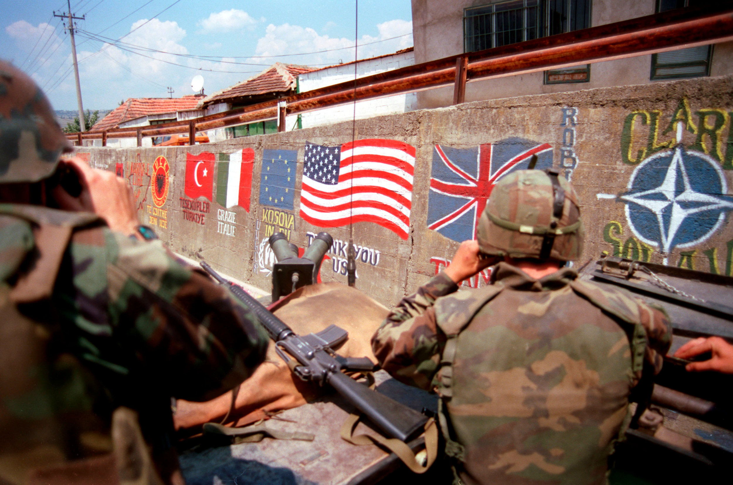 U.S. Marines from the 2nd Light Armored Reconnaissance Battalion pass flags of the NATO nations and messages of thanks painted on the retaining wall of a road in a village in Kosovo on July 5, 1999. (SGT Craig/DOD Photo)