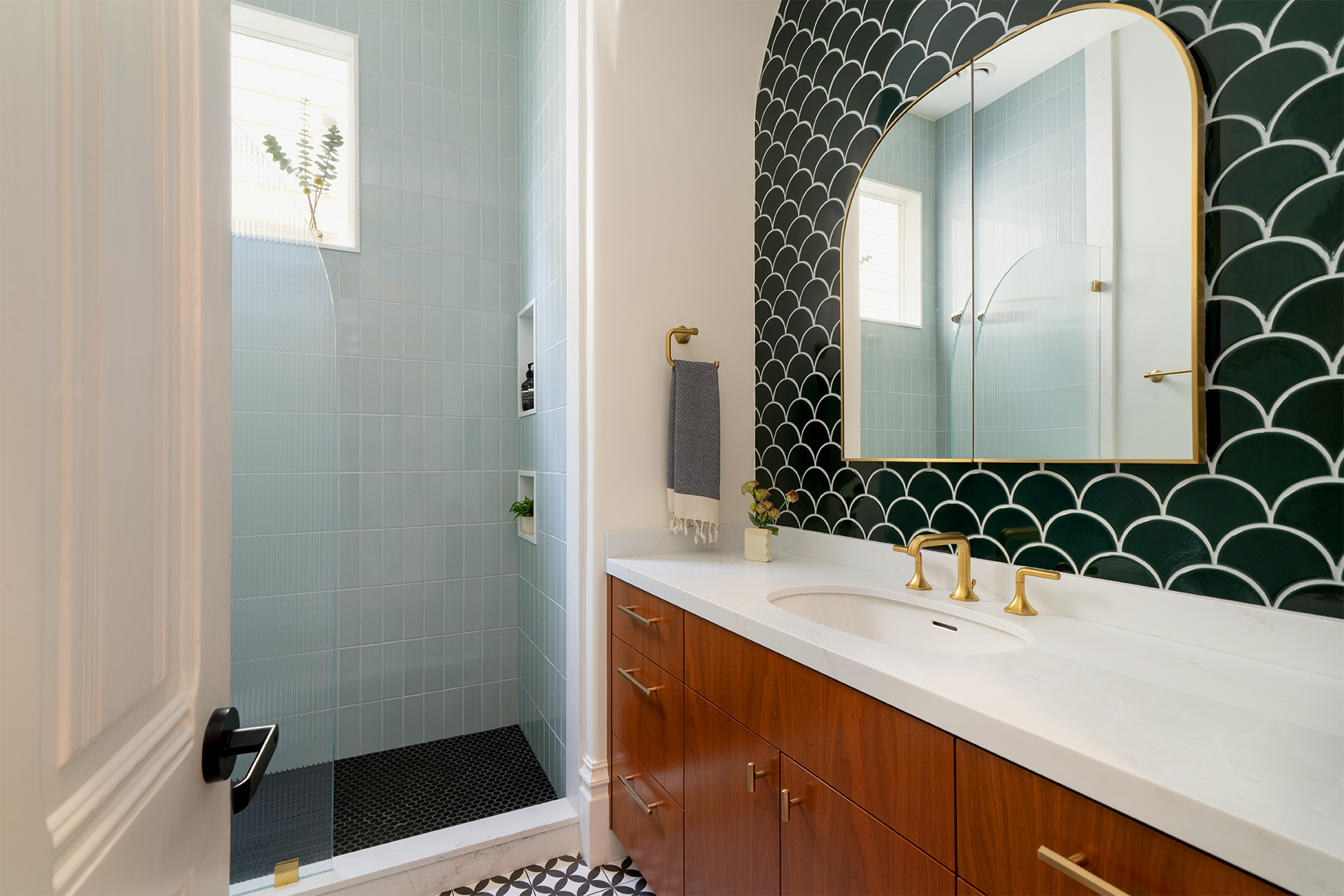 A colorful San Francisco bathroom remodel featuring a custom wood vanity, deep green scalloped tile, pale blue tiled shower, brass fixtures, patterned flooring, and an arched mirror.