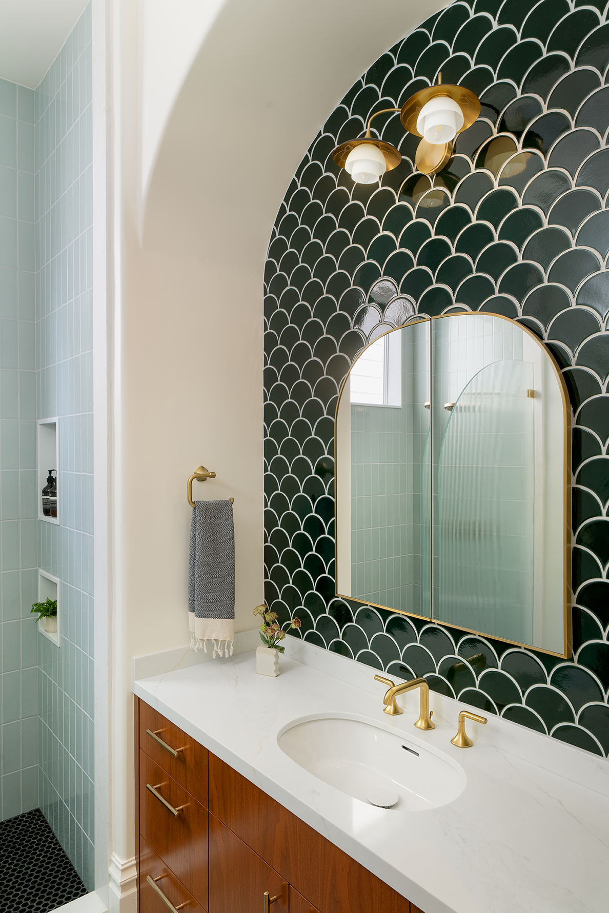 A custom bathroom vanity framed by deep green scalloped tile, an arched brass mirror, warm wood cabinetry, brass fixtures, and a contrasting pale blue tiled shower.