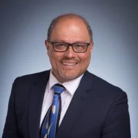 Professional headshot of a man in a suit and glasses, smiling against a gradient blue background.