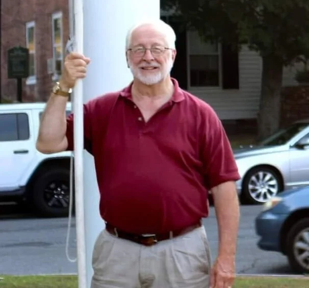 An older man with white hair and glasses smiling, holding a pole outdoors with parked cars and a house in the background.