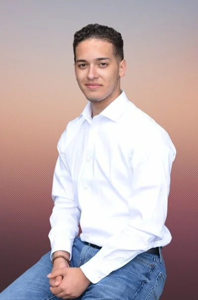 Young man with curly hair wearing a white shirt and blue jeans, sitting against a gradient background.