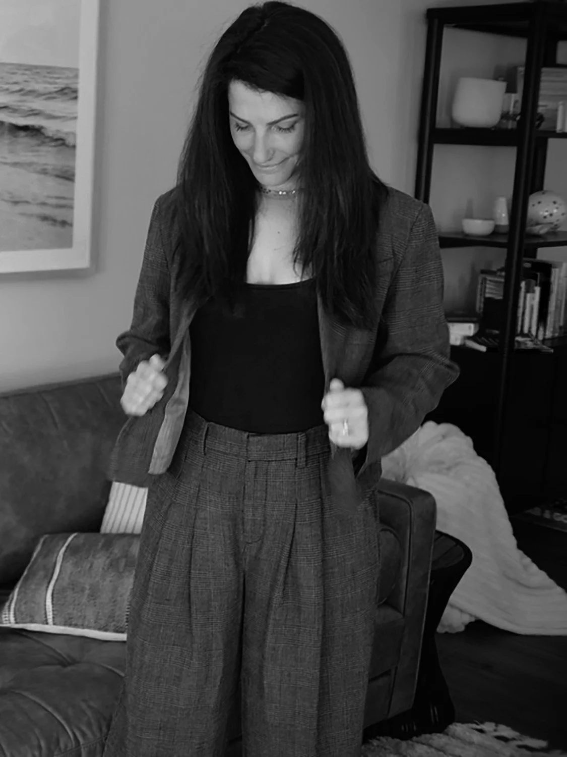 A woman with long dark hair is smiling and looking down, wearing a plaid suit jacket and matching pants, with a black top underneath, in a living room setting.