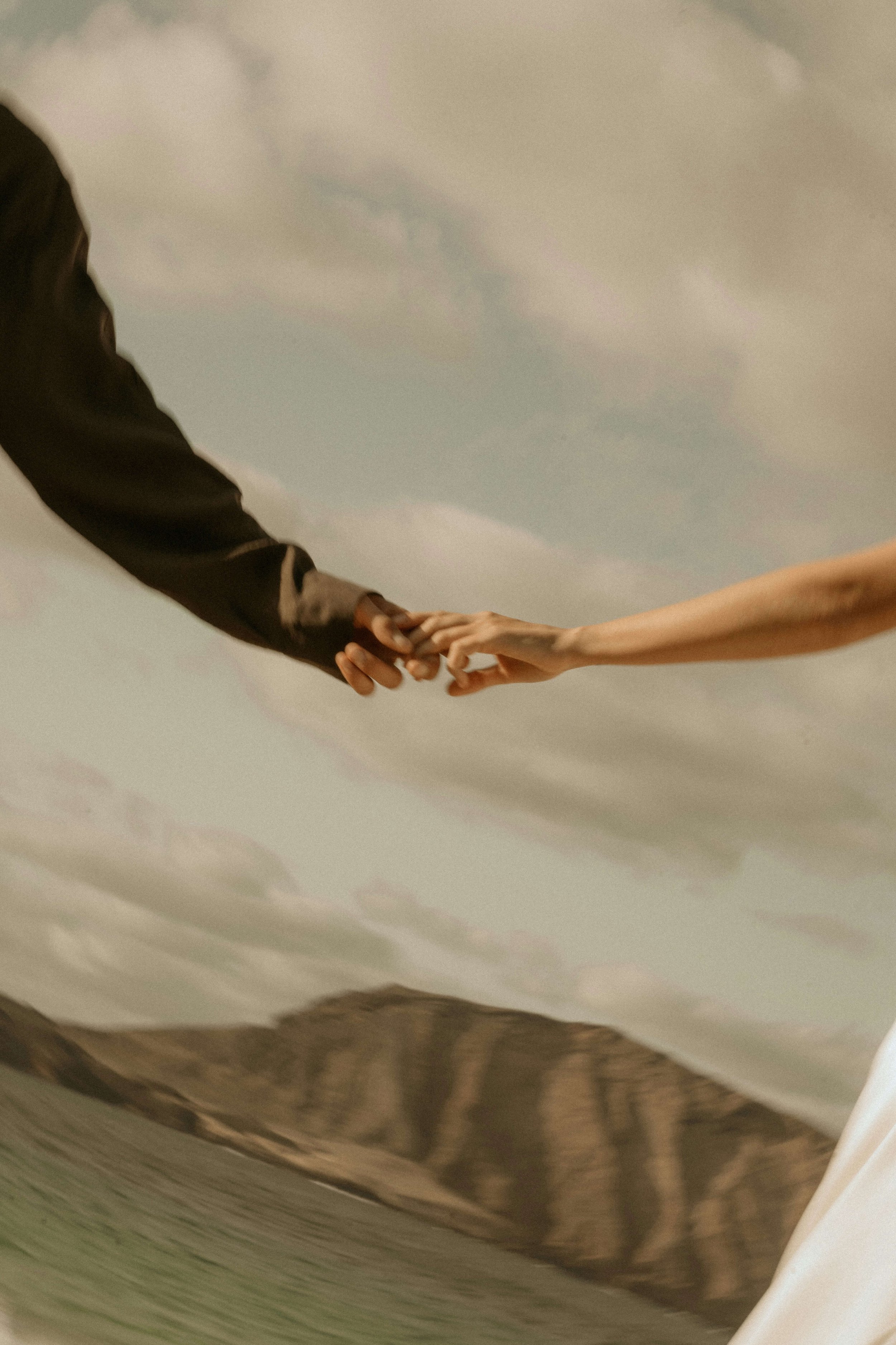 A close-up image of two hands, one masculine with a darker skin tone and one feminine with a lighter skin tone, reaching towards each other with fingers almost touching, against a neutral background.