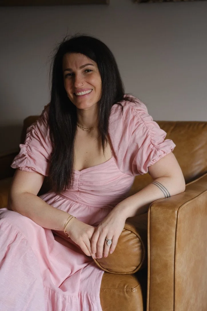 Woman with long dark hair in a pink dress sitting on a brown leather couch, smiling at the camera.