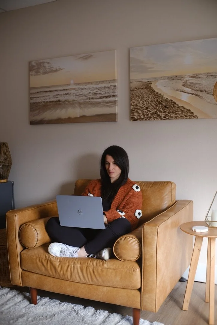 Woman sitting on a tan leather couch working on a laptop in a living room, with beach-themed landscape paintings on the wall behind her.