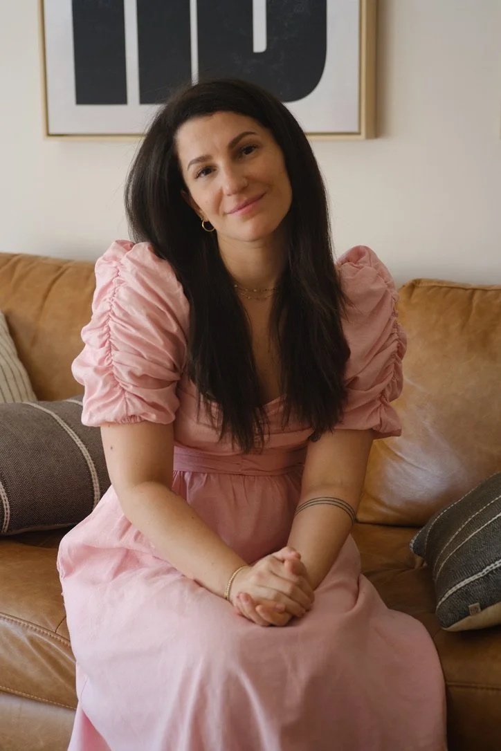 A woman with long dark hair, wearing a pink dress with puffed sleeves, sits on a tan couch with striped pillows, smiling and clasping her hands together.