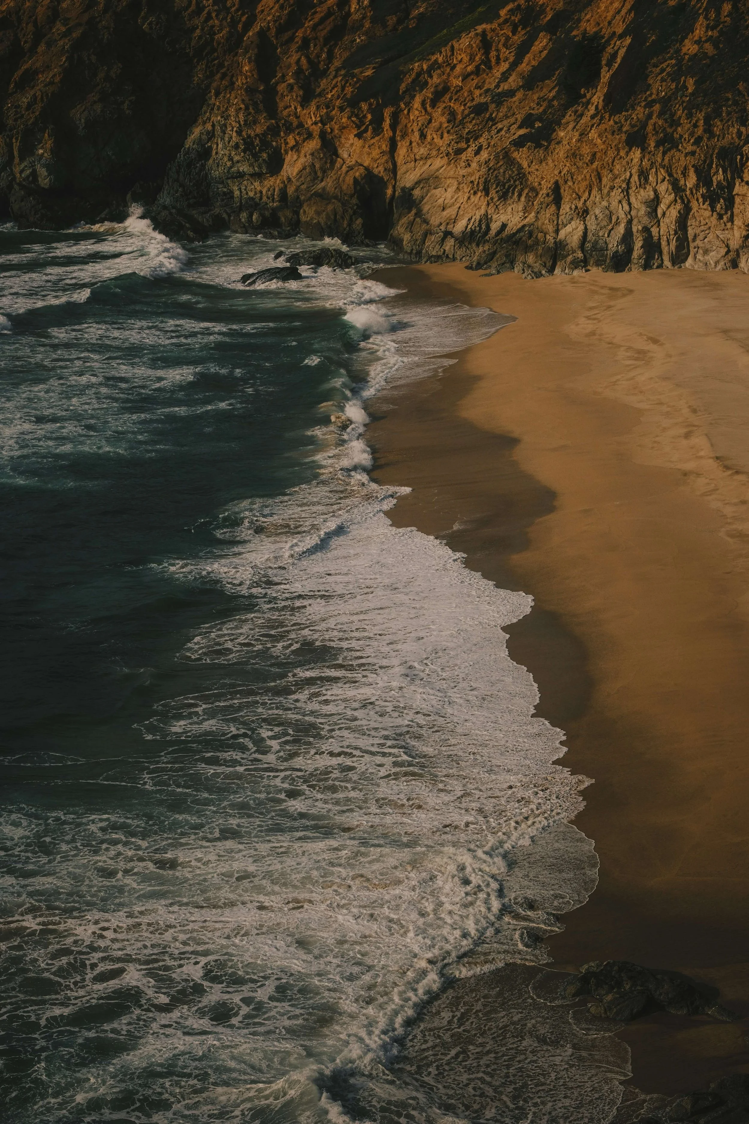 A rugged coastline with steep cliffs and rocky outcroppings, waves crashing onto a sandy beach at sunset.